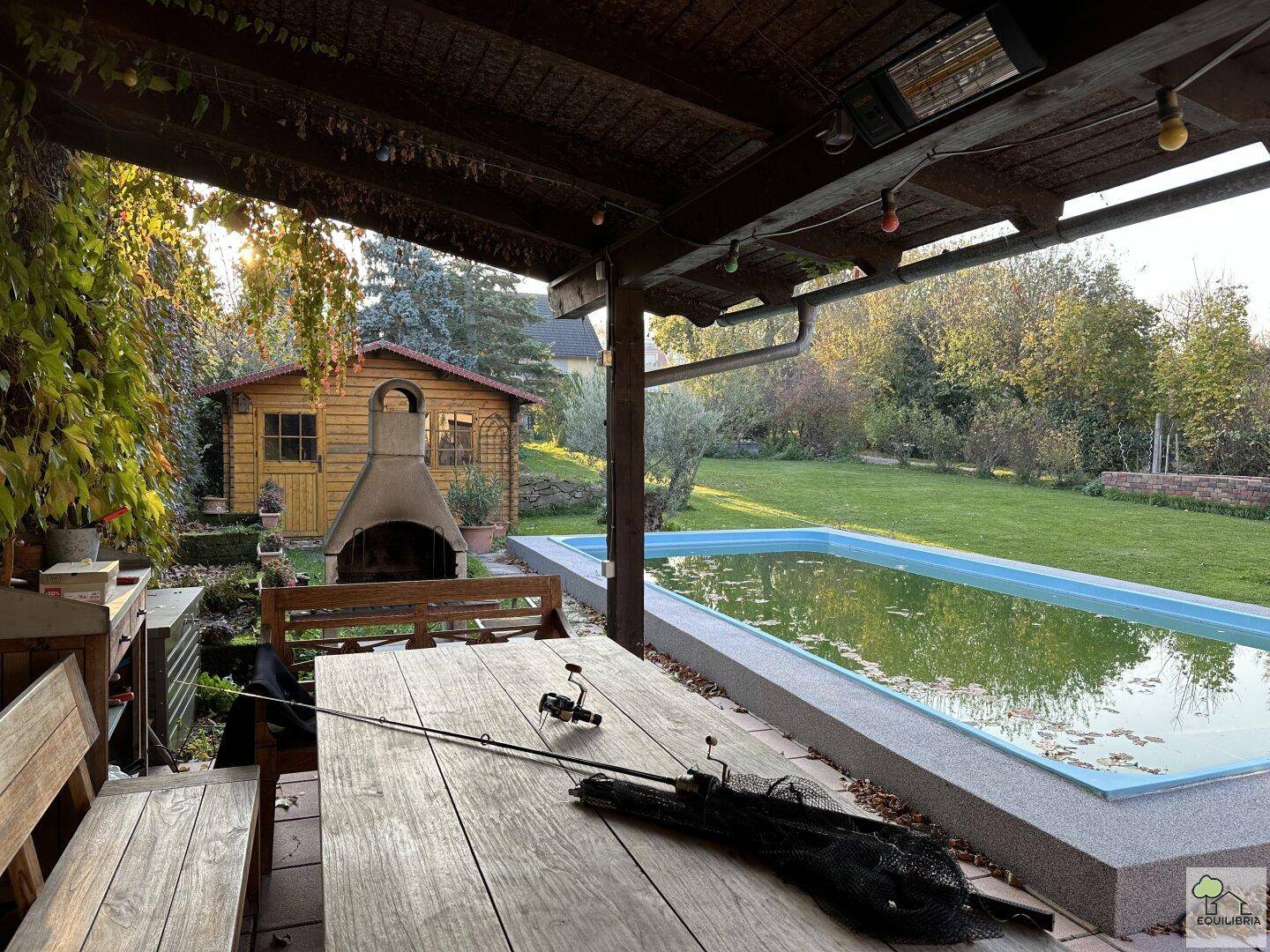 Überdachter Außenbereich mit Holztisch und Bänken, Blick auf Pool und Garten.