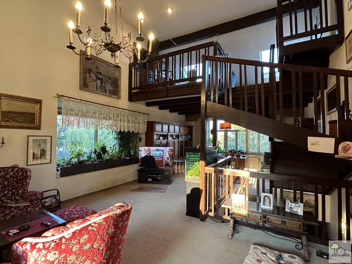 Geräumiges Wohnzimmer mit hoher Decke, großer Holztreppe und traditioneller Einrichtung.
