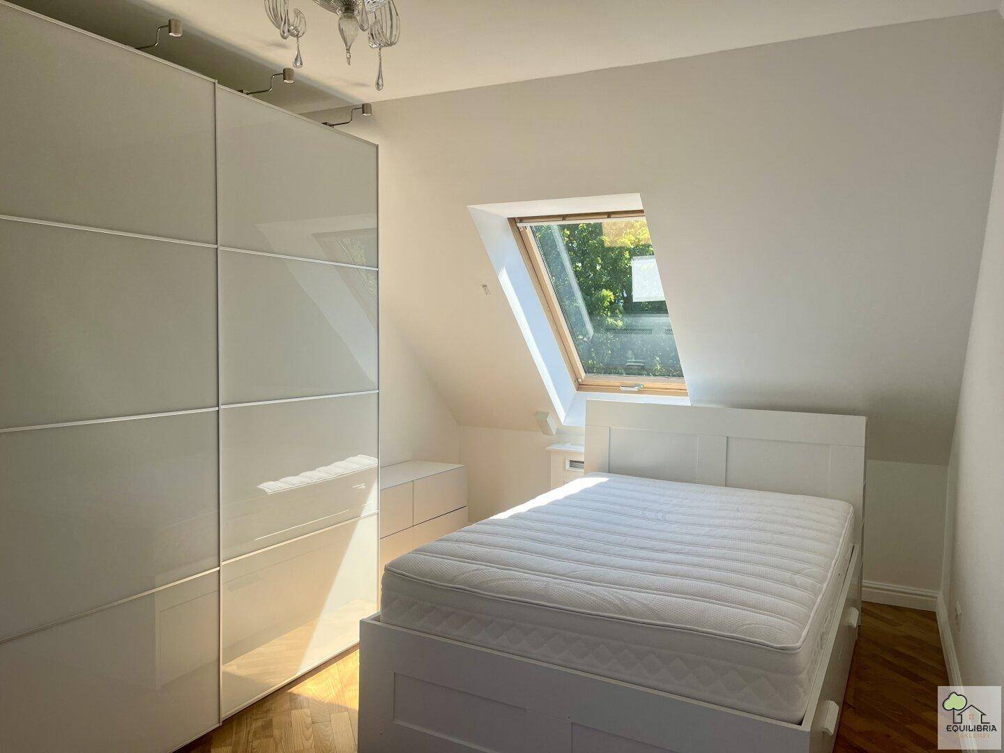Gemütliches Schlafzimmer mit weißem Bett, Einbauschrank und schrägem Dachfenster für viel Licht.