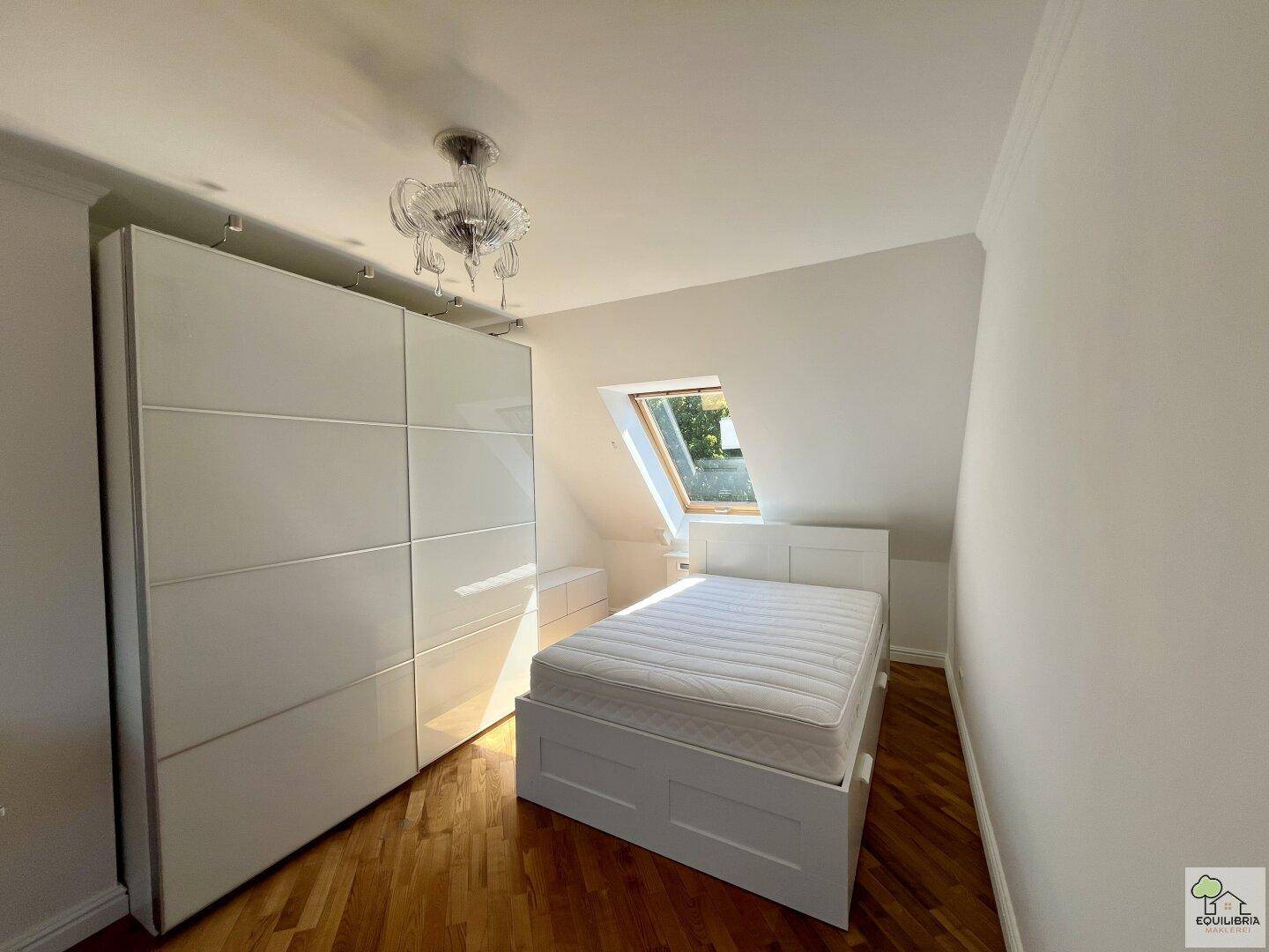 Helles Schlafzimmer mit weißem Bett, großem Kleiderschrank und einem Dachfenster.