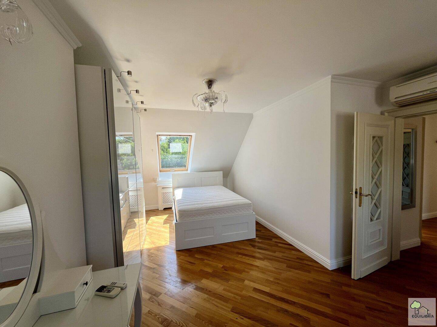 Schlafzimmer mit weißem Bett, Dachfenster, Schminktisch und Klimaanlage.