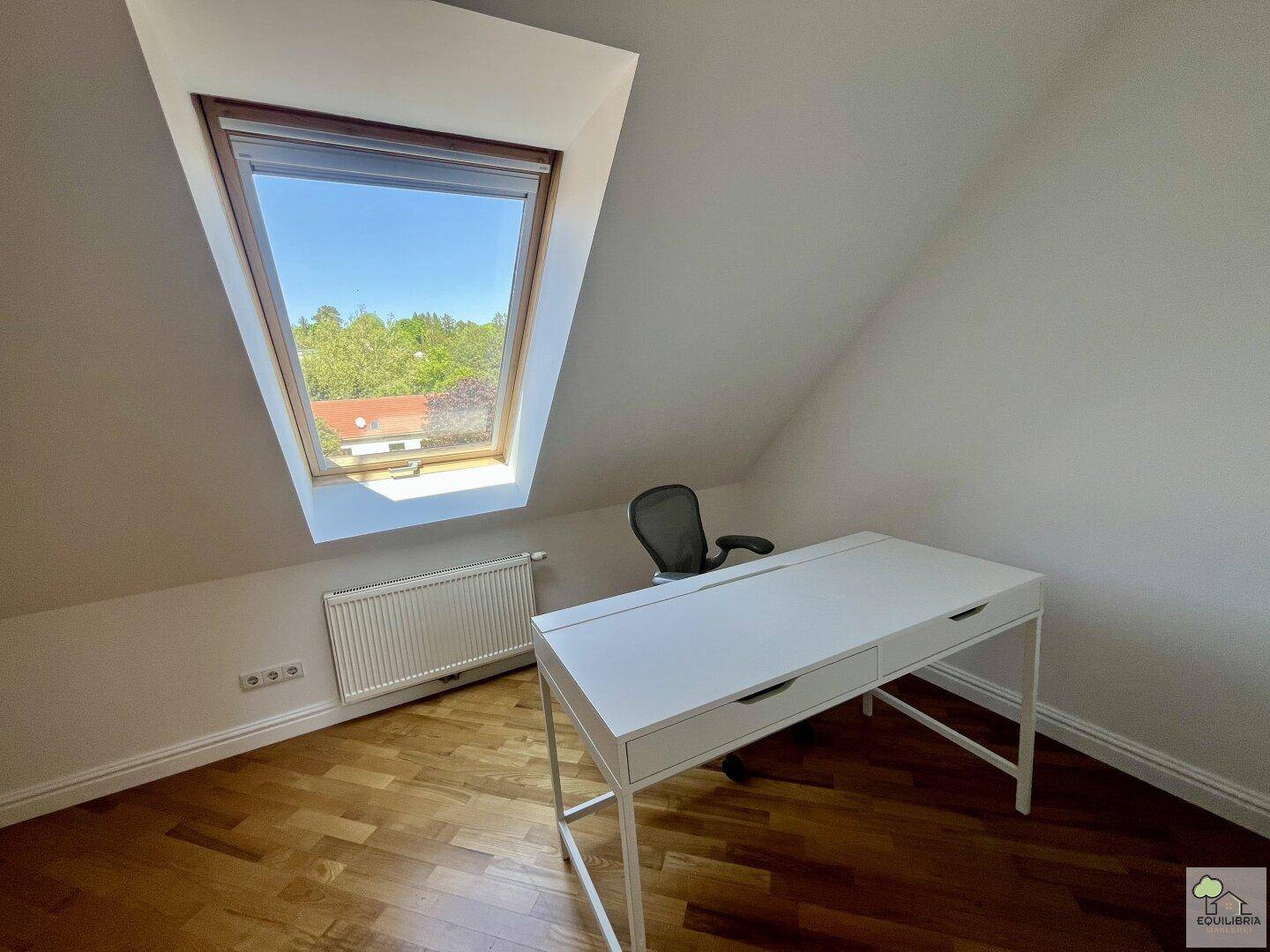 Ruhiger Arbeitsplatz mit Dachfenster, weißem Schreibtisch und Blick ins Grüne.