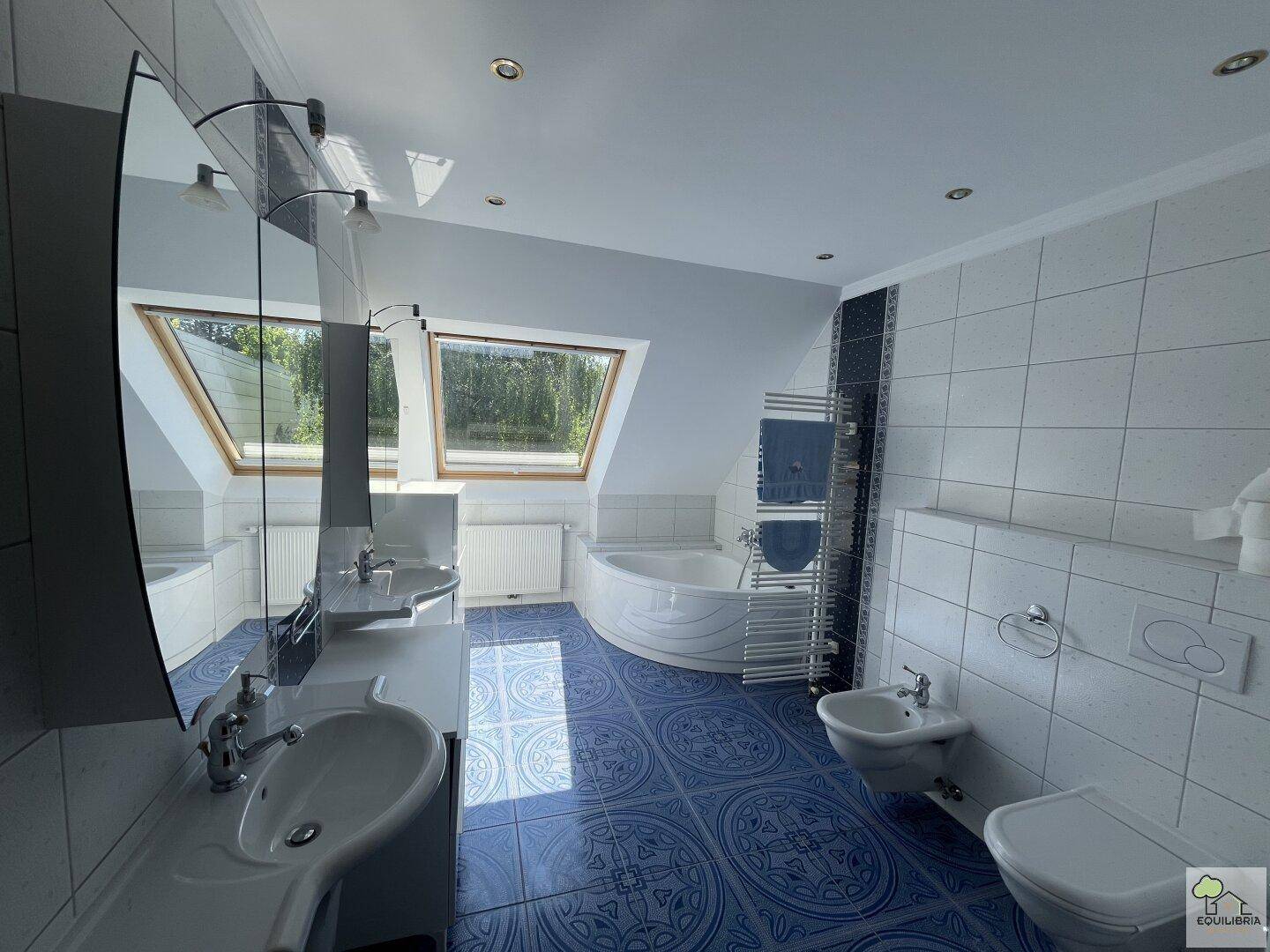 Helles Badezimmer mit blauen Fliesen, Doppelwaschbecken, Eckbadewanne und Dachfenstern.