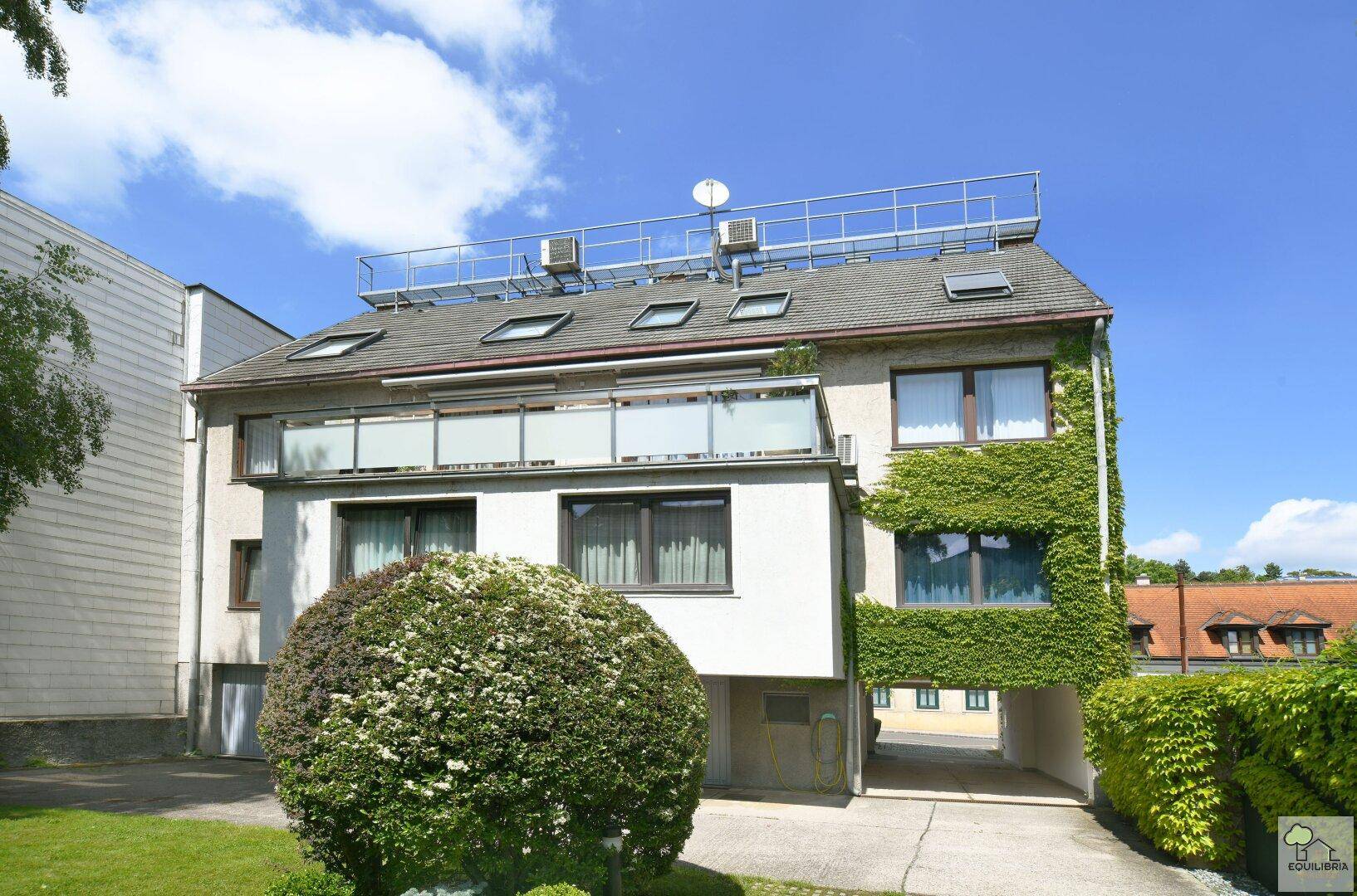 Mehrstöckiges Gebäude mit begrüntem Gartenbereich, Balkon und einer Dachterrasse.