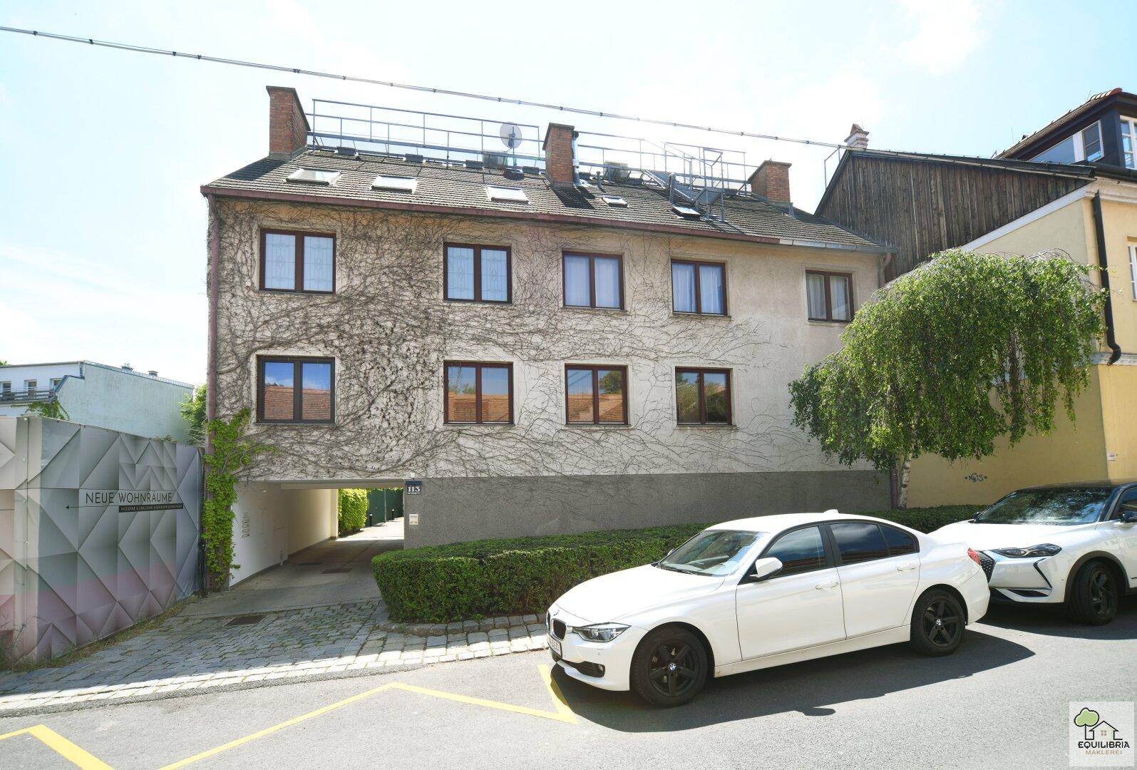 Fassade eines Gebäudes mit Efeubewuchs, Parkplätzen und einem Durchgang.