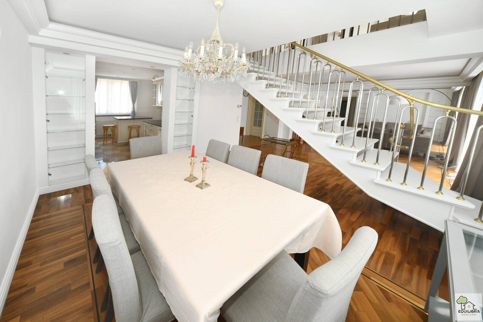 Eleganter Essbereich mit großem Tisch, Kronleuchter und Zugang zu einer geschwungenen Treppe.