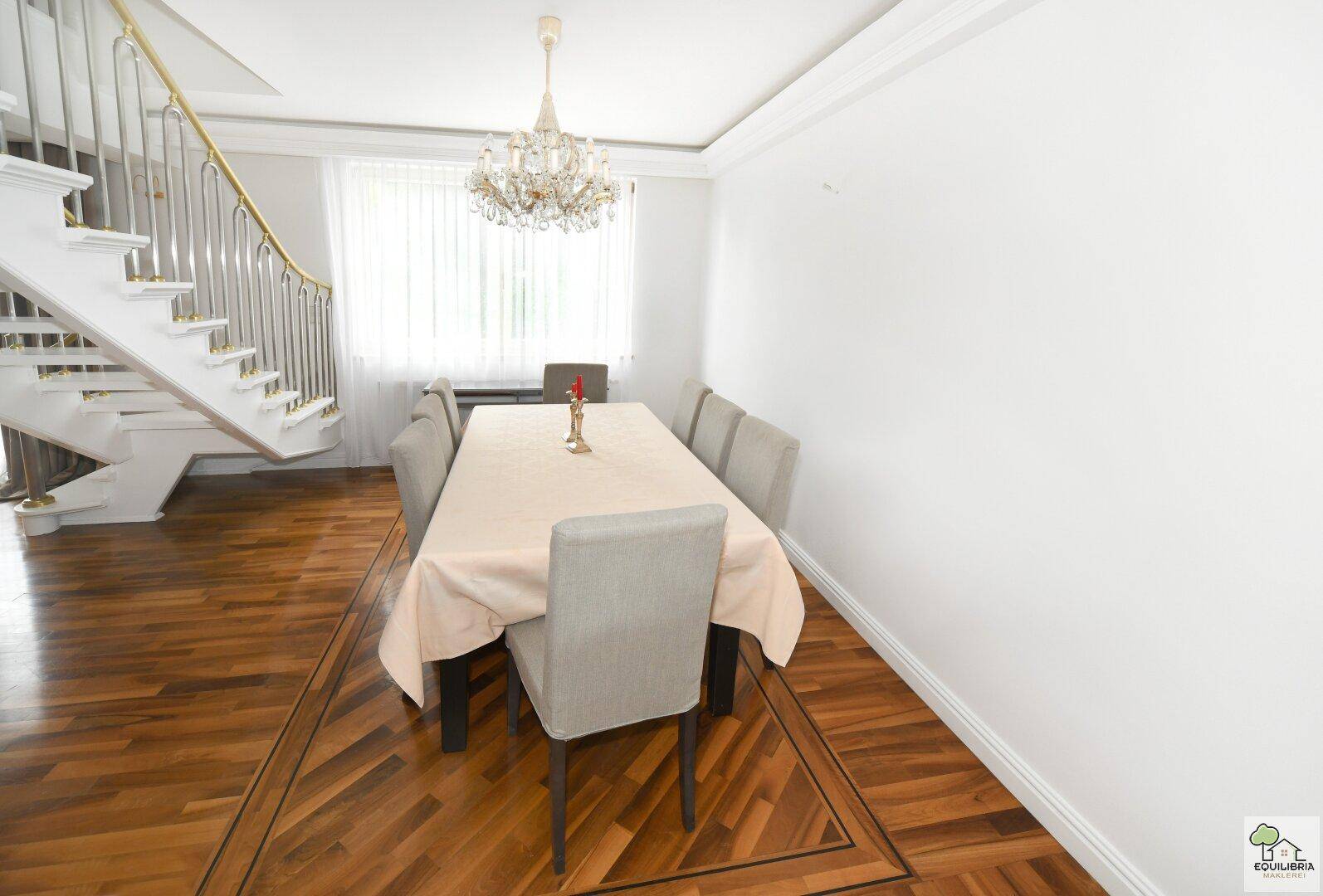 Eleganter Essbereich mit großem Tisch, Kronleuchter und Zugang zu einer geschwungenen Treppe.