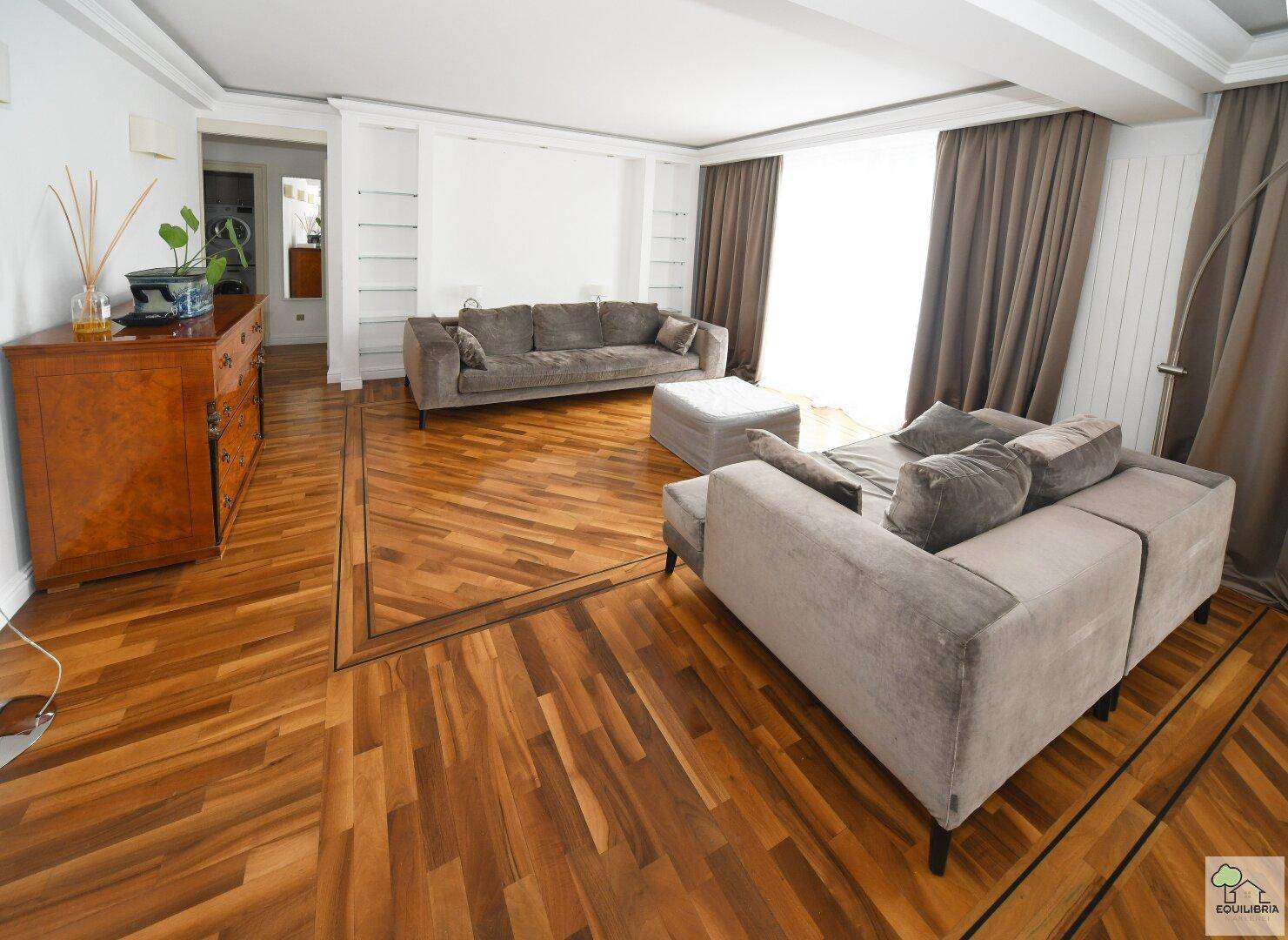 Helles Wohnzimmer mit Parkettboden, bequemen Sofas und großen Fenstern mit braunen Vorhängen.