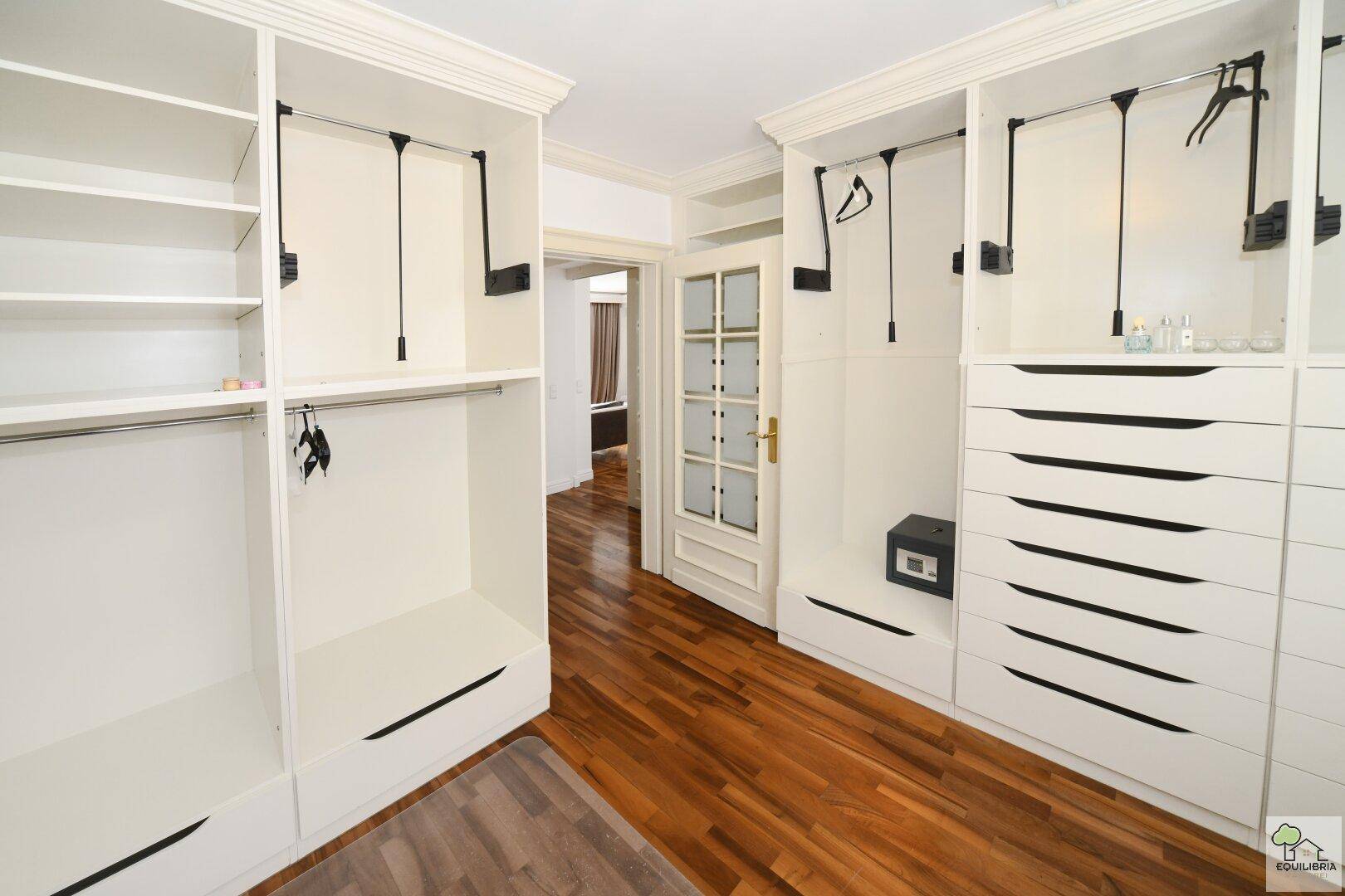 Begehbarer Kleiderschrank mit weißen Einbauschränken und Zugang zu einem weiteren Raum.