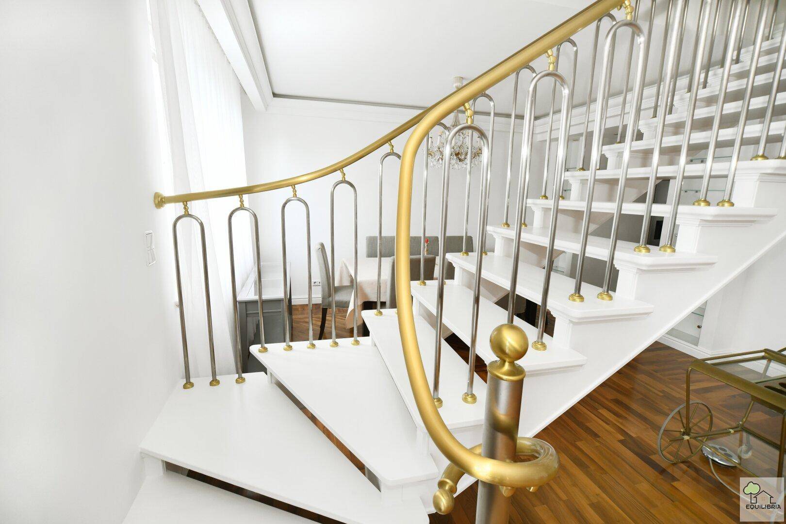 Detailansicht einer eleganten Treppe mit goldenem Handlauf und weißen Stufen.