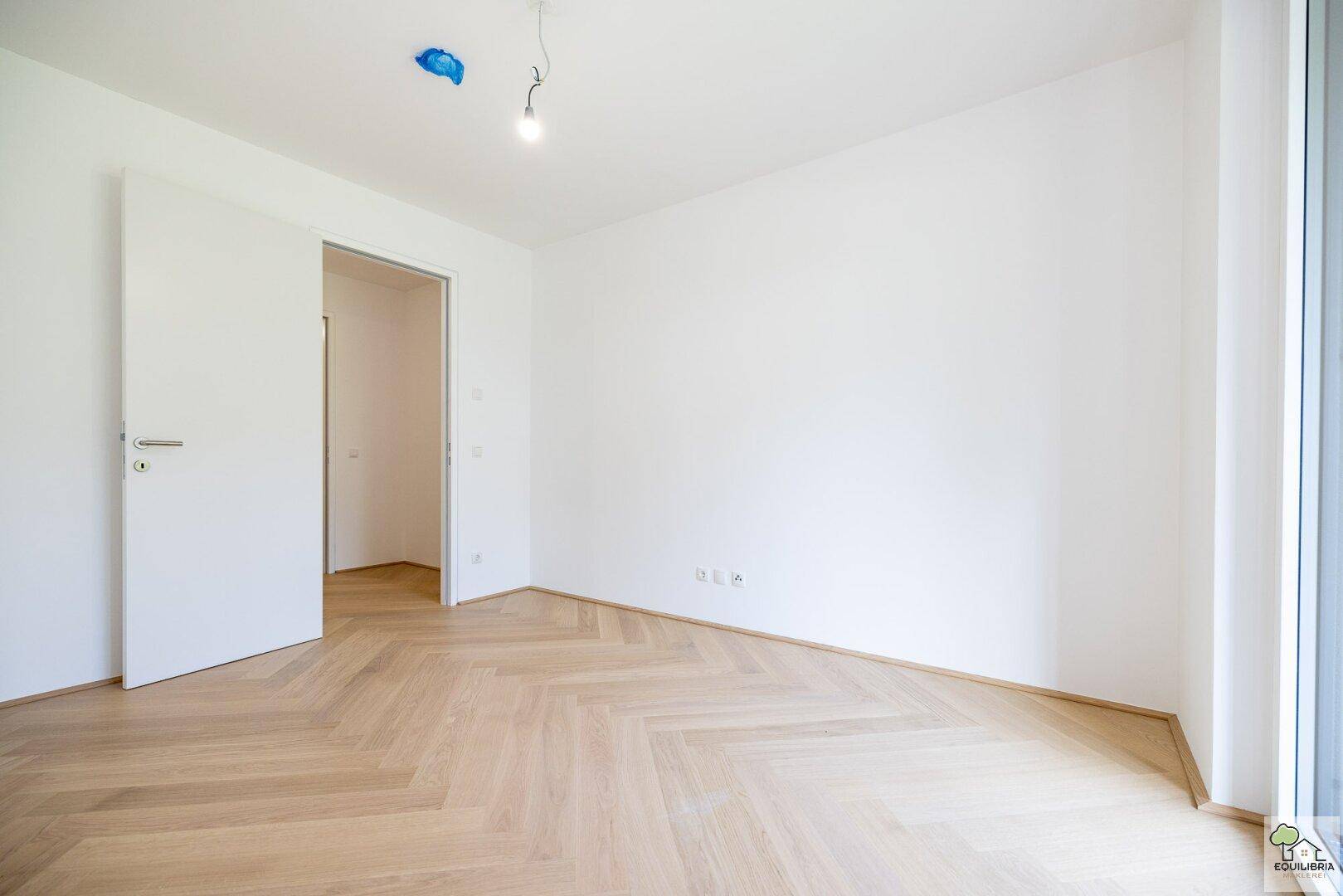 Geräumiges, leeres Zimmer mit hellem Fischgrätparkett und weiß gestrichenen Wänden.
