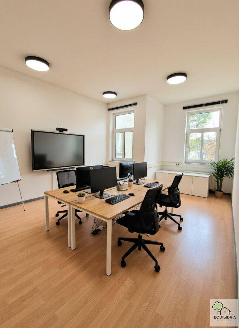 Modern eingerichtetes Büro mit mehreren Arbeitsplätzen, großem Bildschirm und Fenstern mit Tageslicht.