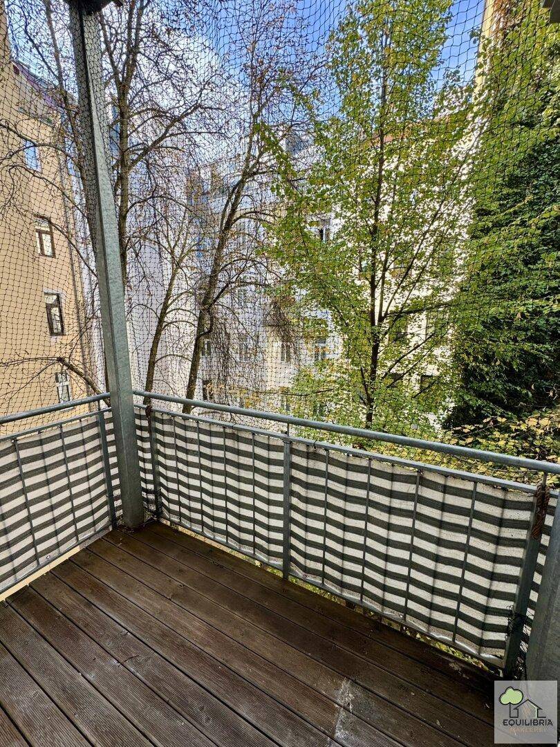 Kleiner Balkon mit Holzboden und Blick auf Bäume sowie umliegende Gebäude im Innenhof.