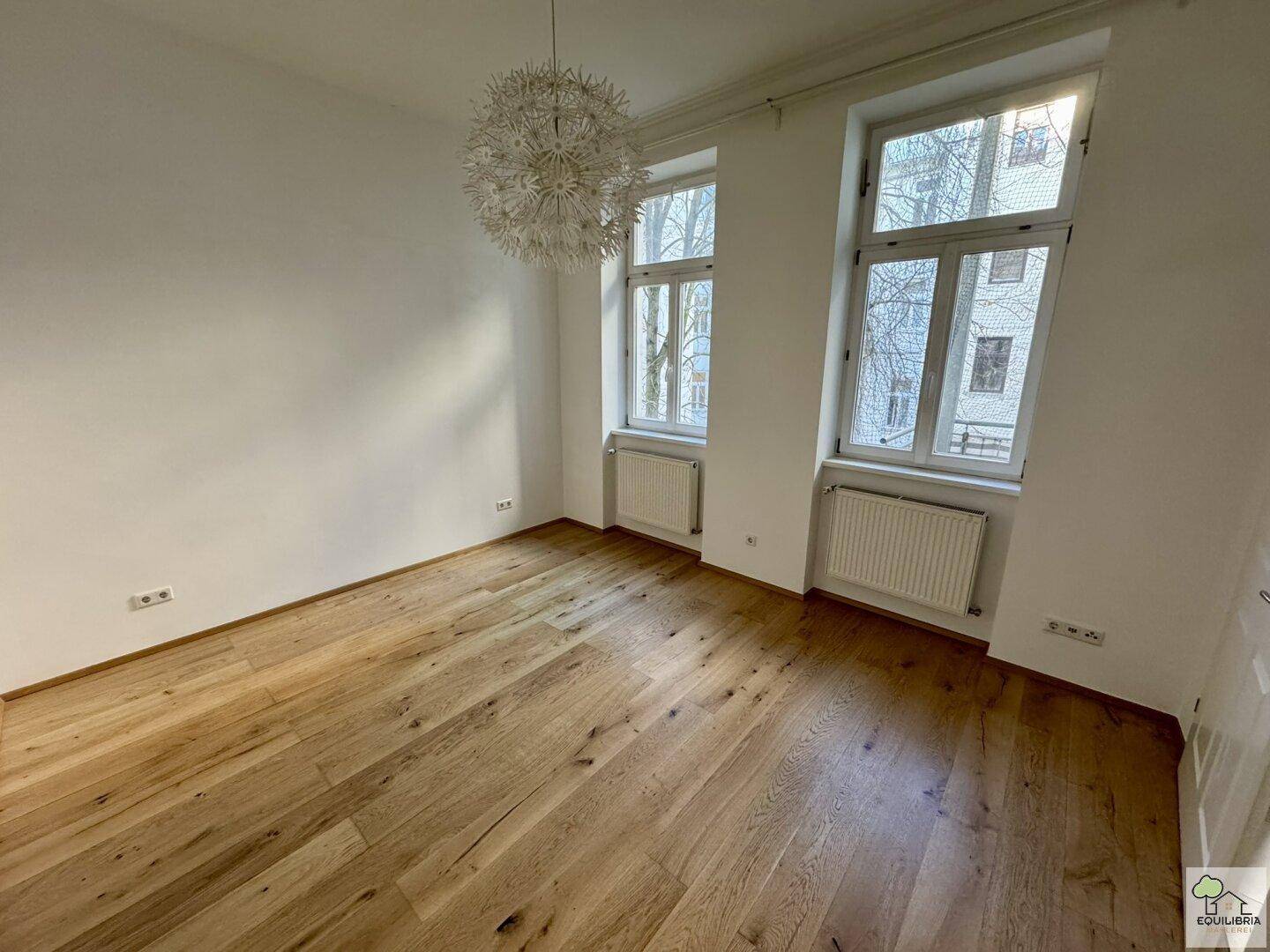 Geräumiges Zimmer mit Holzparkett, hohen Decken und zwei großen Fenstern mit Blick nach draußen.