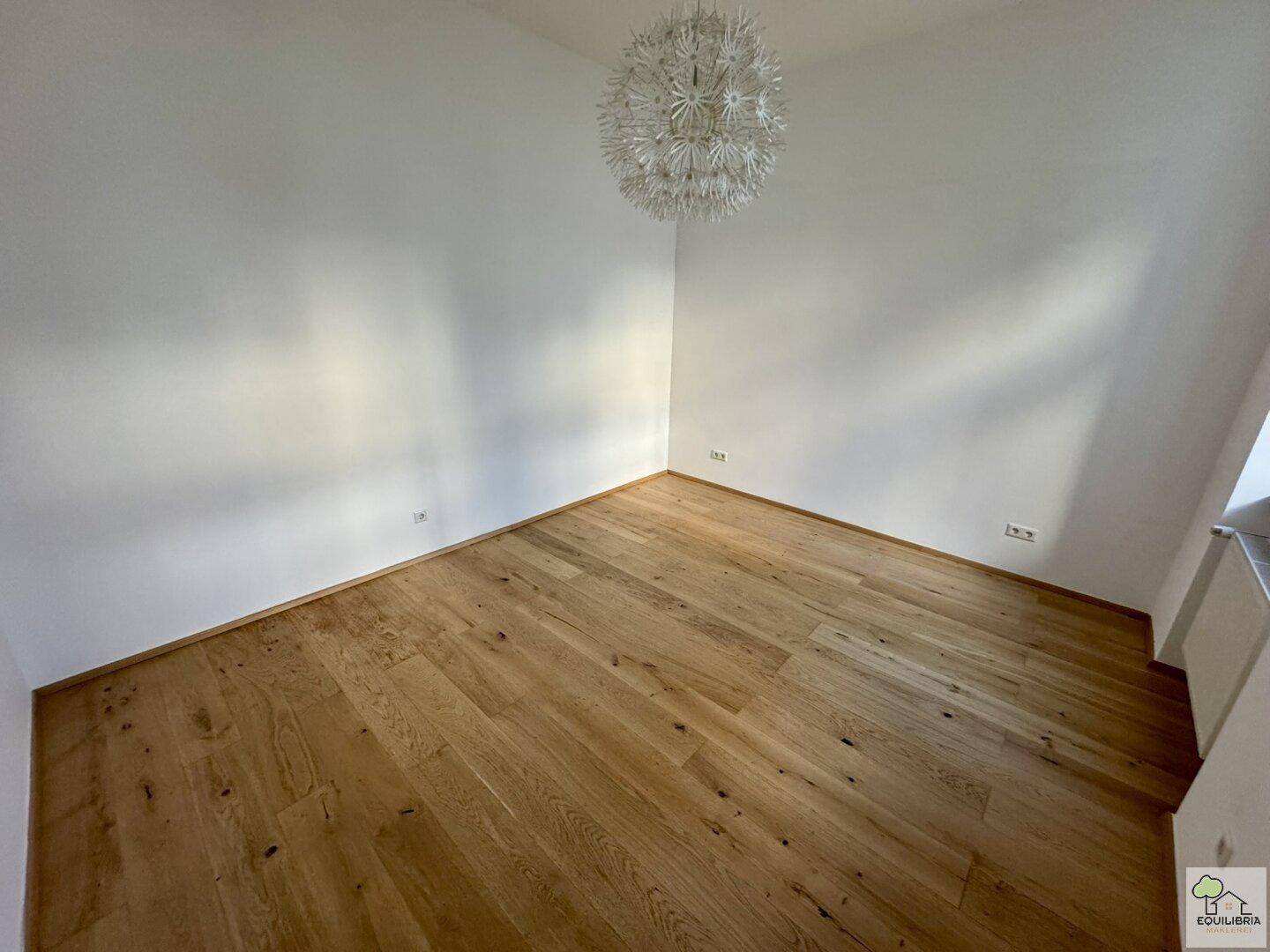 Helles Zimmer mit Holzparkettboden, weißen Wänden und einer modernen Deckenleuchte.