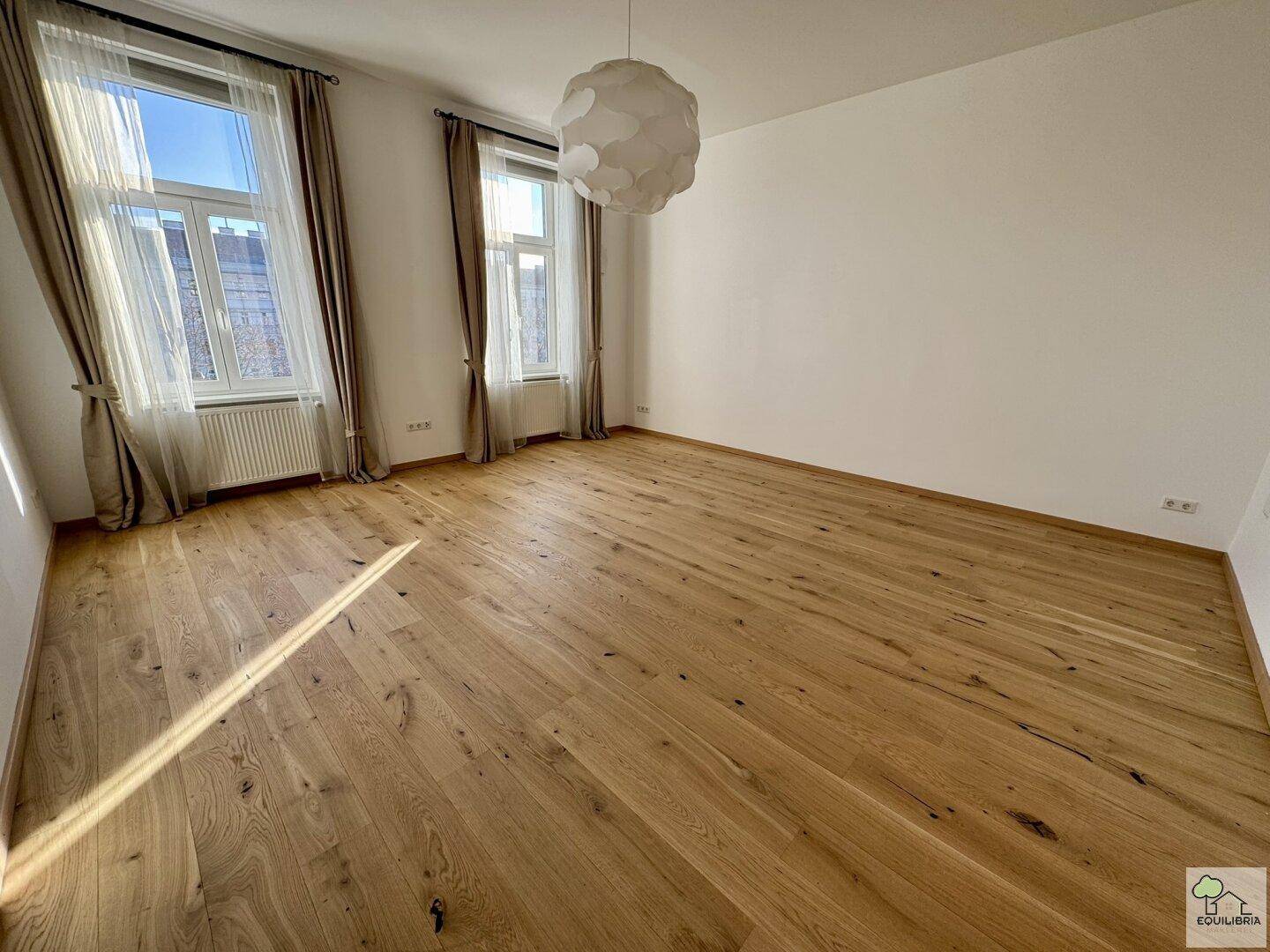 Lichtdurchflutetes Zimmer mit hellem Holzparkett und viel Sonneneinstrahlung durch die großen Fenster.