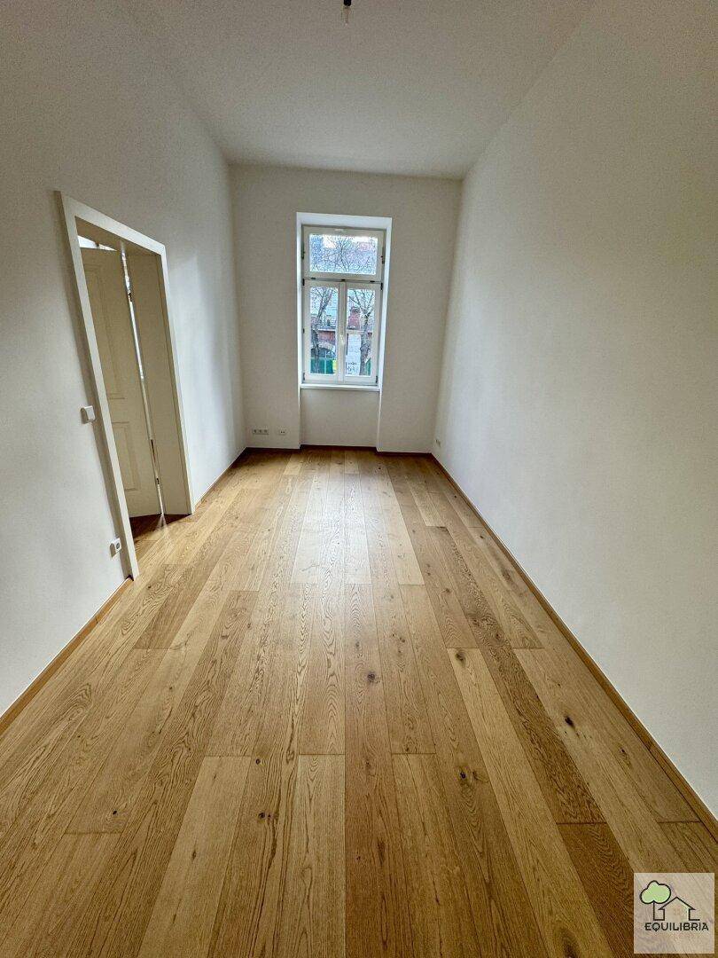 Langes, helles Zimmer mit Holzboden und einem Fenster, ideal für ein Kinder- oder Arbeitszimmer.