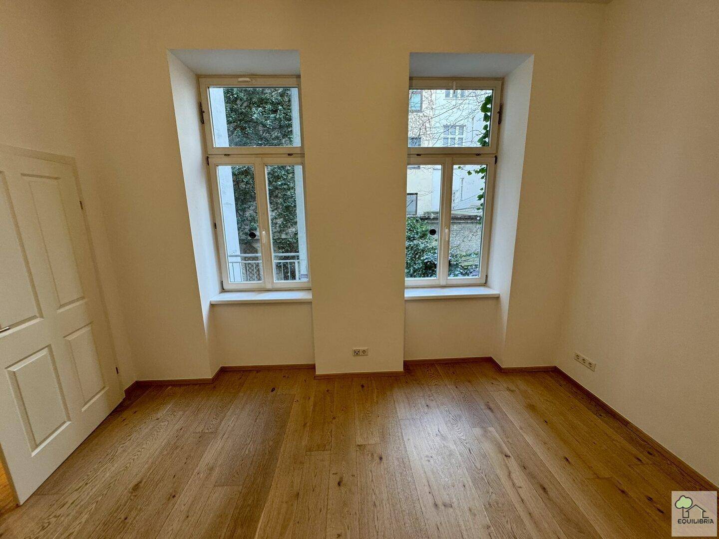 Helles Zimmer mit Holzboden und zwei hohen Fenstern, geeignet als Schlaf- oder Arbeitszimmer.