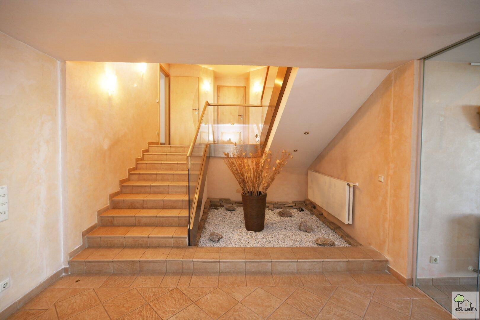 Heller Flur mit gefliester Treppe, Glasgeländer und dekorativem Kiesbeet.