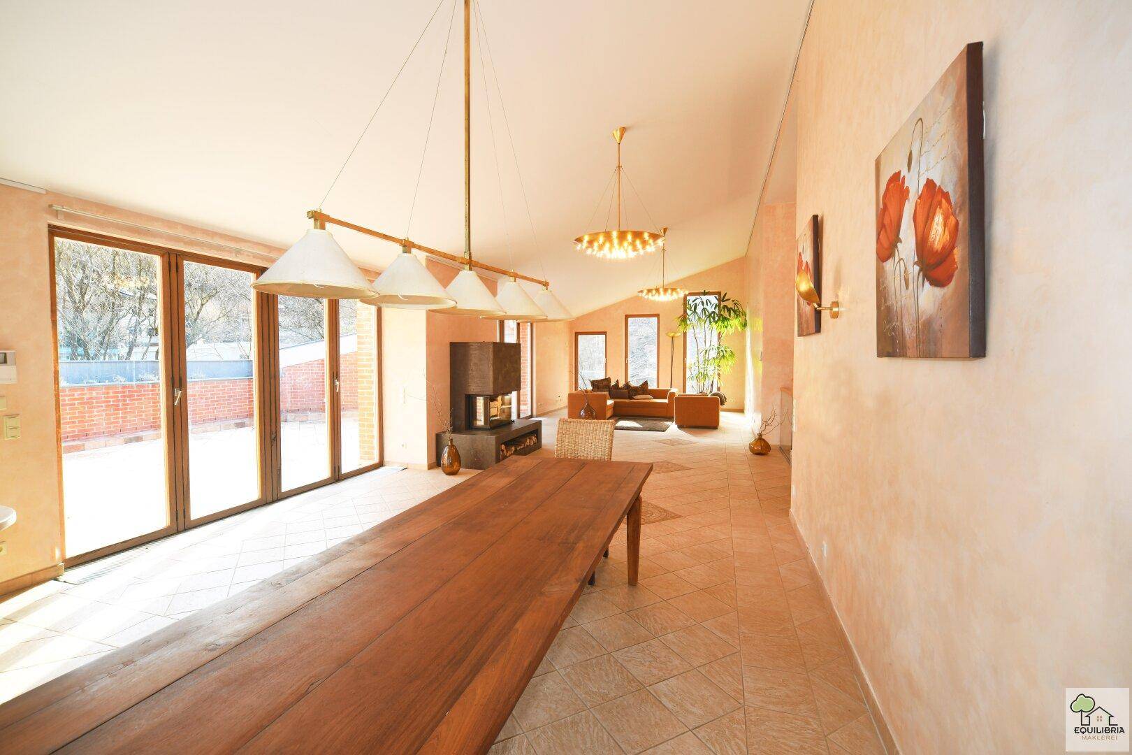 Geräumiger Wohnbereich mit langem Esstisch, Kamin und großen Fensterfronten zur Terrasse.