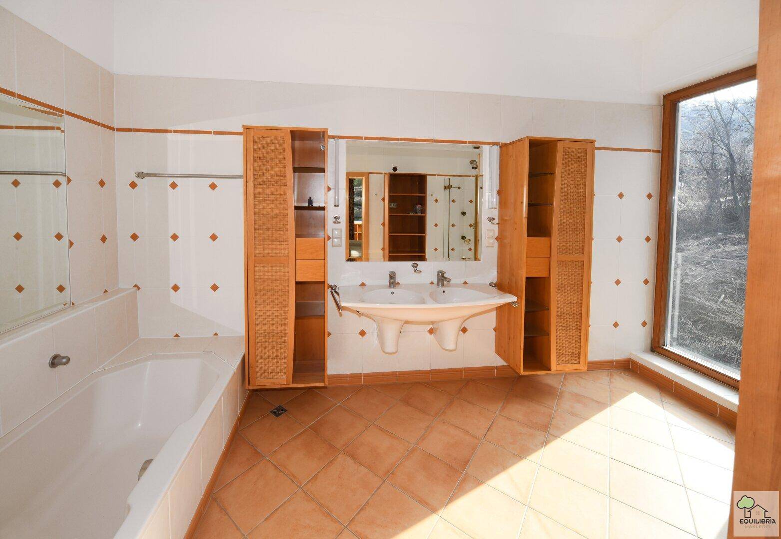 Helles Badezimmer mit Badewanne, Doppelwaschbecken und großen Fenstern.