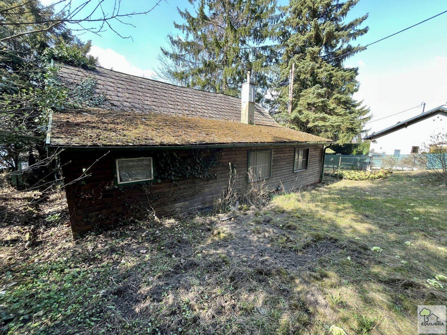 Rustikales Holzgebäude mit Moos bewachsenem Dach, umgeben von trockenem Gras und Bäumen.