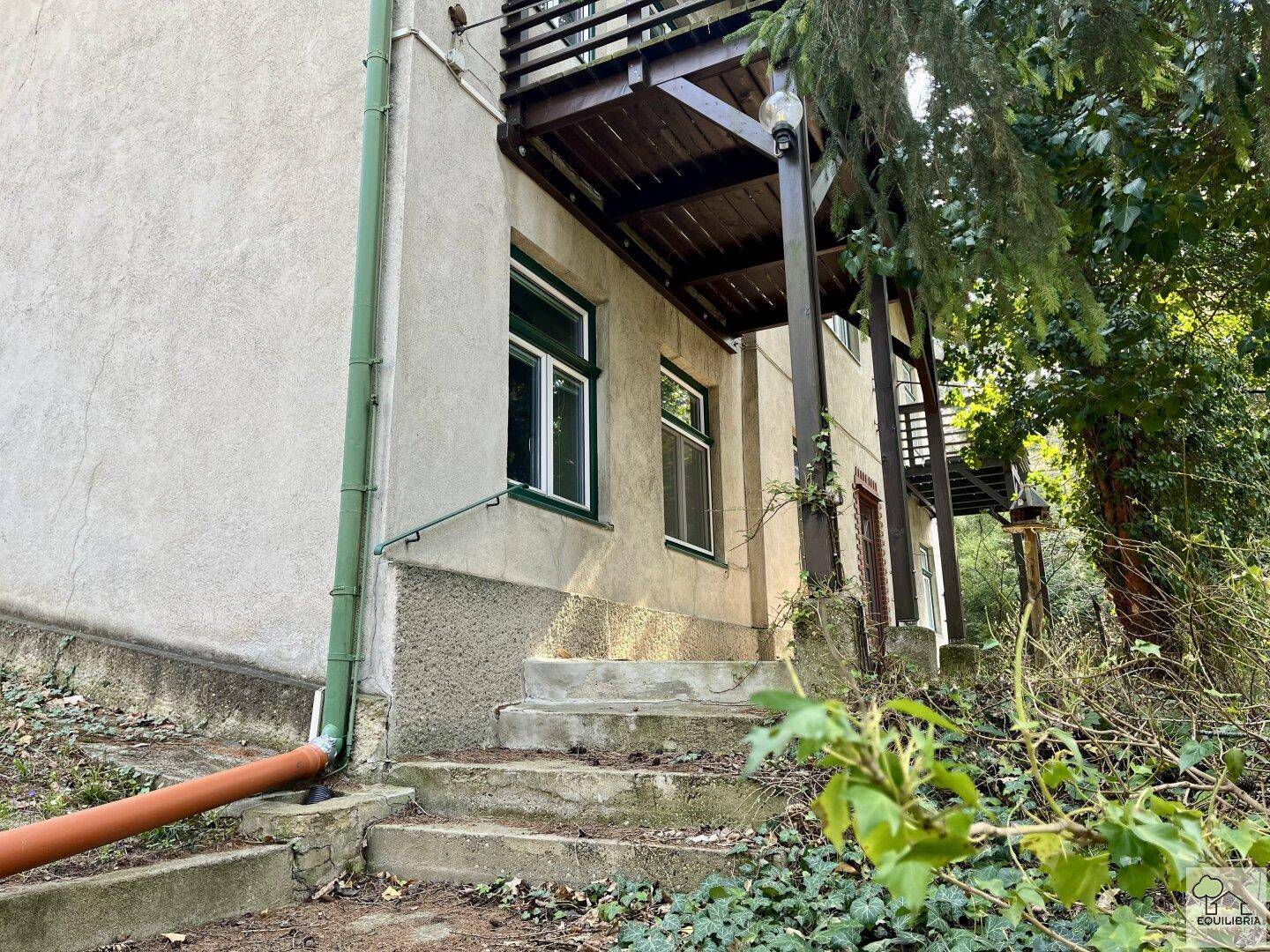 Seitenansicht des Hauses mit Außentreppe und einem Balkon, umgeben von Vegetation.