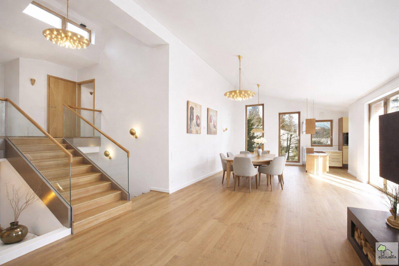Offener Wohnbereich mit Essplatz, Holztreppe und viel Tageslicht durch hohe Fenster.