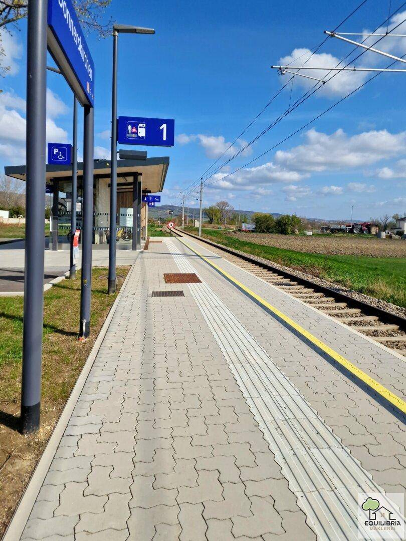 Moderner Bahnsteig mit Beschilderung und Gleisen, umgeben von grünen Feldern unter blauem Himmel.