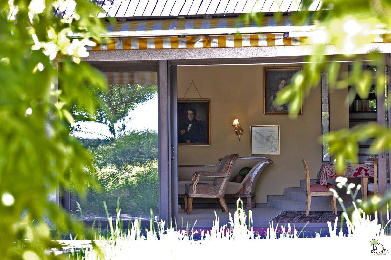 Blick von außen in einen hellen Wohnbereich mit klassischen Möbeln und Zugang zum Garten.