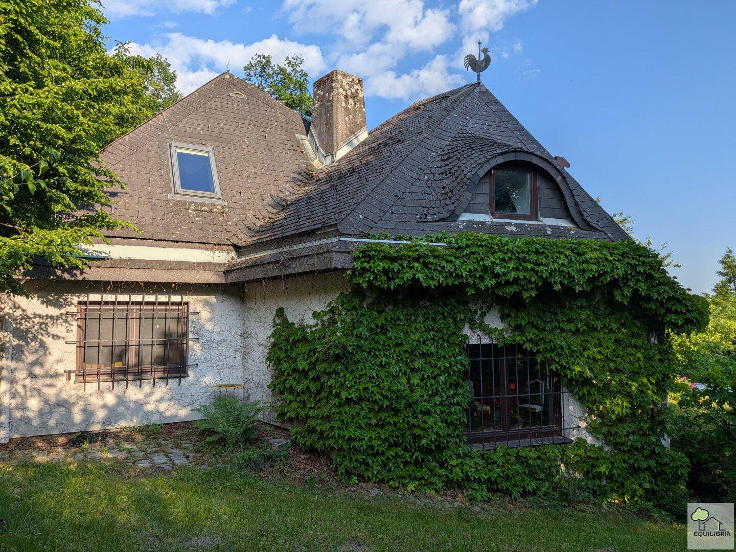 Historisches Haus mit charakteristischem Dach und bewachsenen Fassaden in grüner Umgebung.
