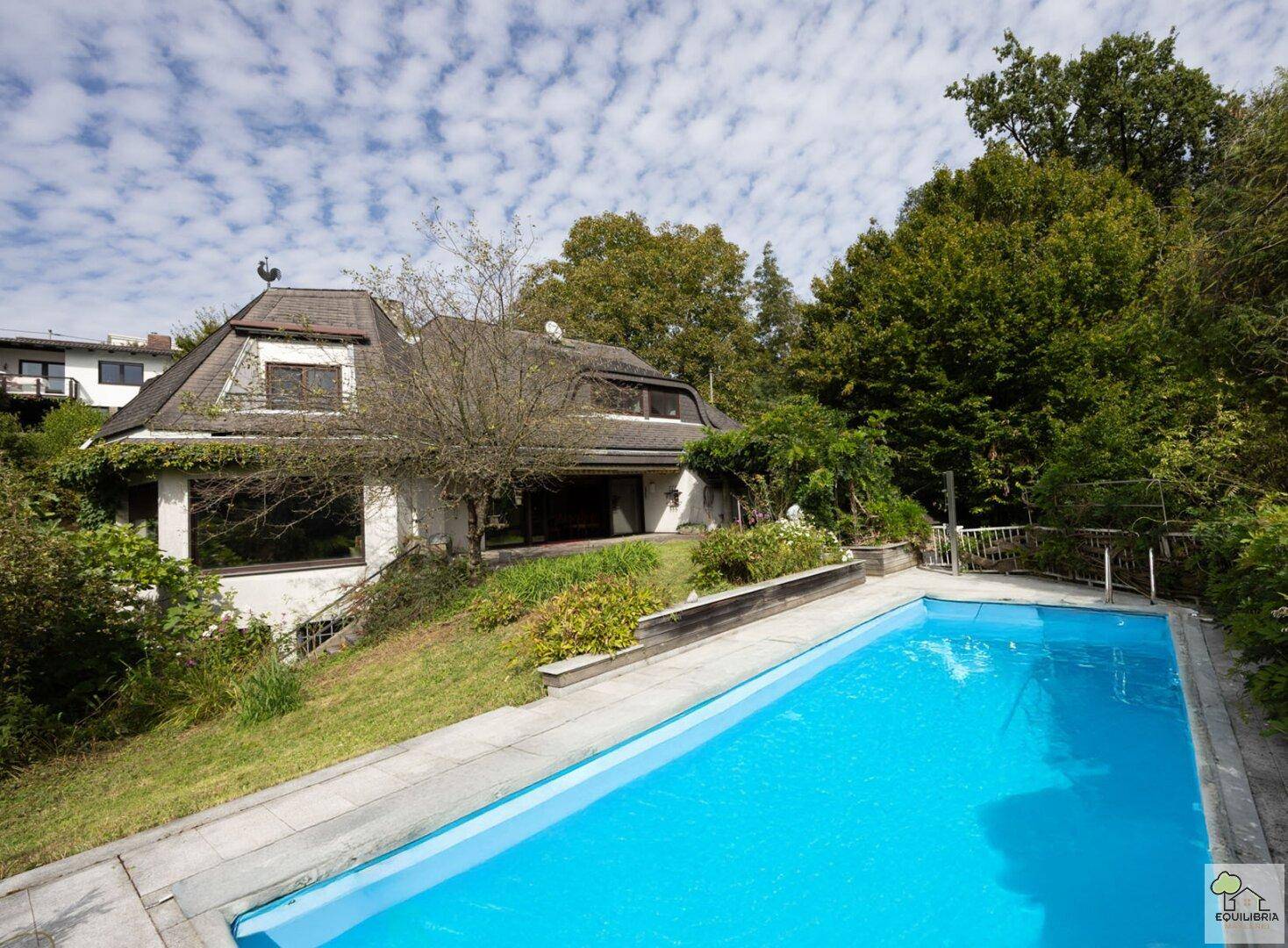Haus mit großem Außenpool, umgeben von viel Grün und einem gepflegten Gartenbereich unter blauem Himmel.