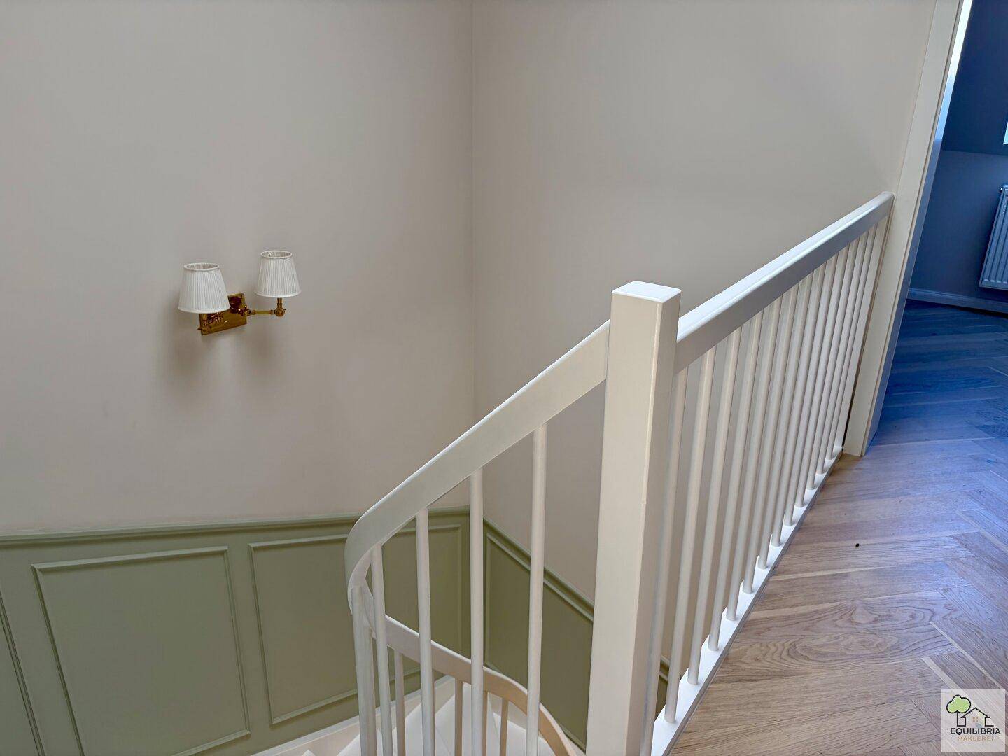 Elegantes Treppenhaus mit weißem Geländer und grüner Wandvertäfelung, führt in die obere Etage.