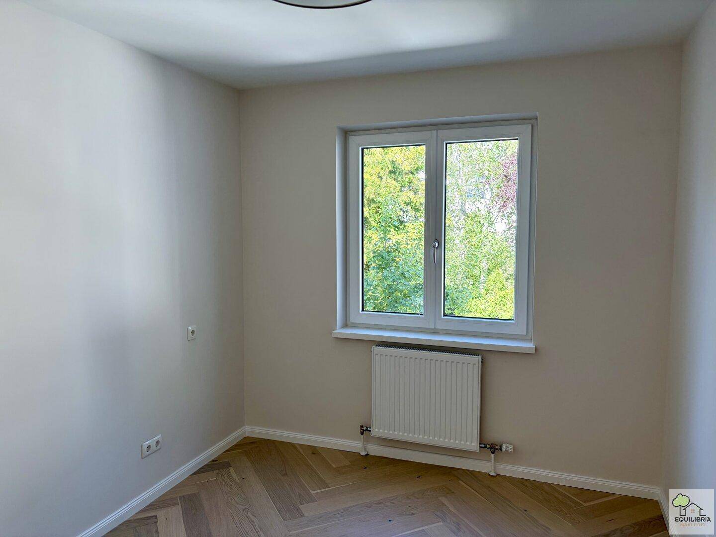 Helles Schlafzimmer mit Fischgrätparkett und großem Fenster mit Blick ins Grüne.