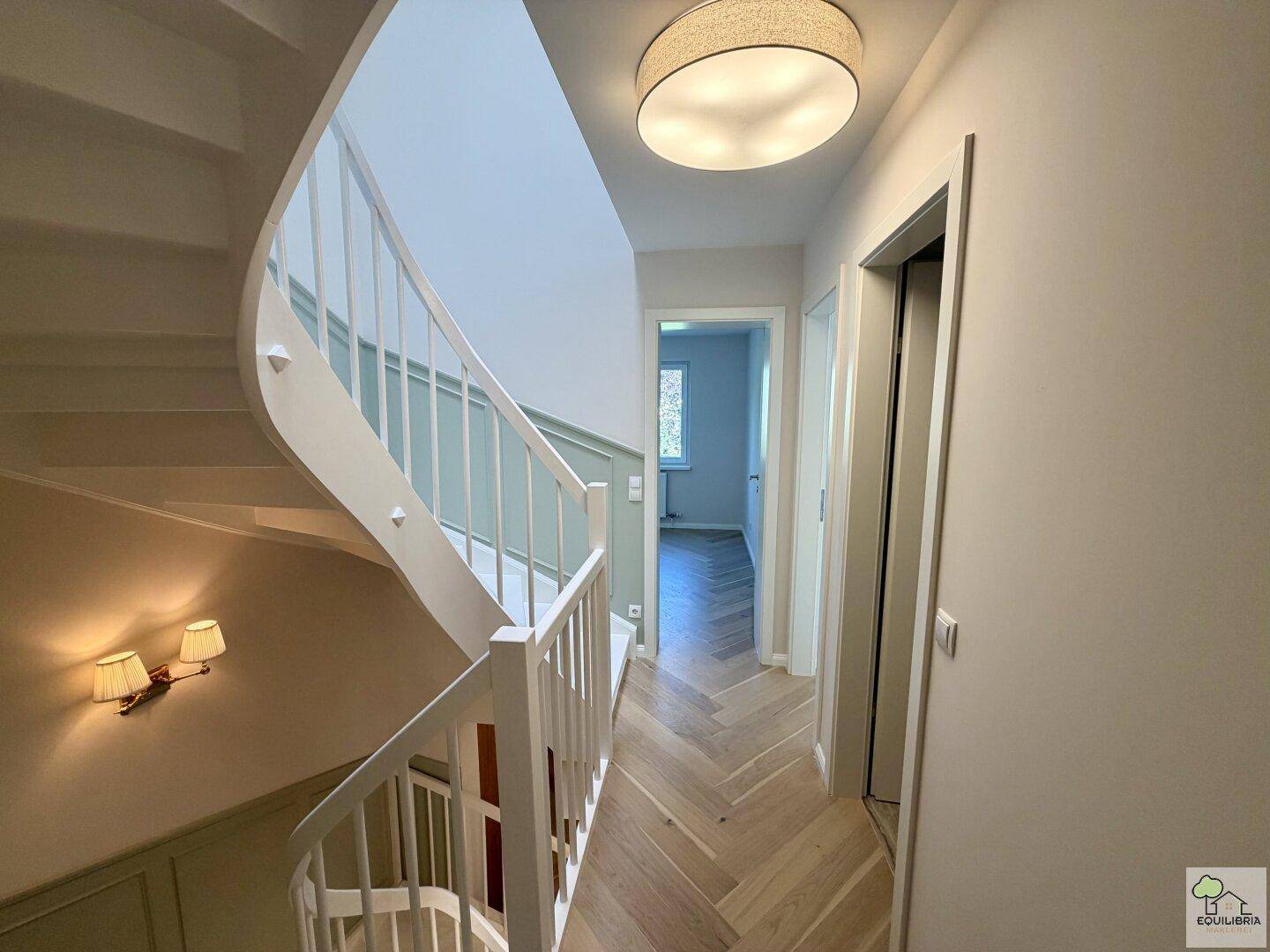 Heller Flur mit geschwungener Treppe und eleganten Wandleuchten, führt zu den Wohnbereichen.