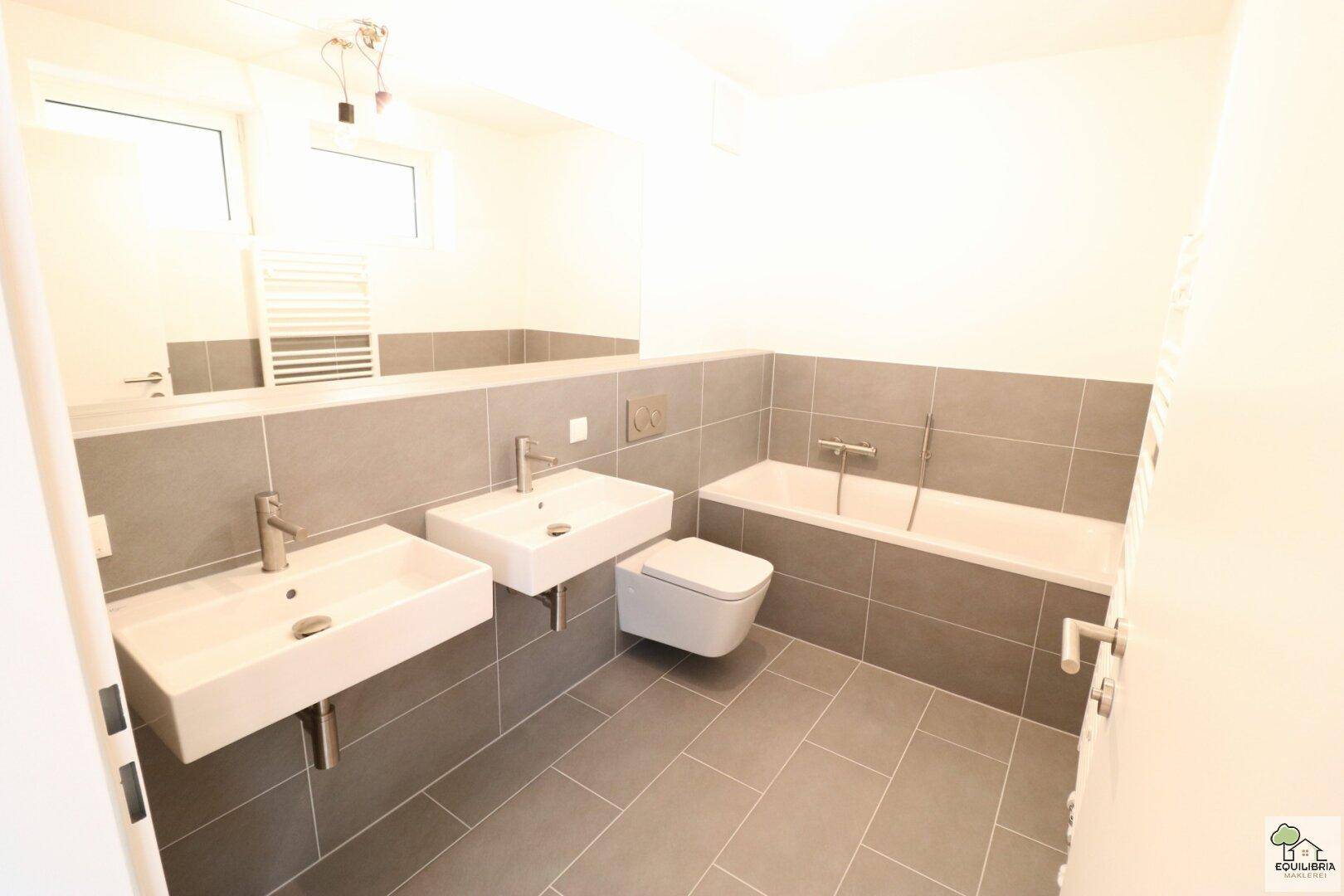 Modernes Badezimmer mit Doppelwaschbecken, Badewanne, wandhängendem WC und grauen Fliesen.
