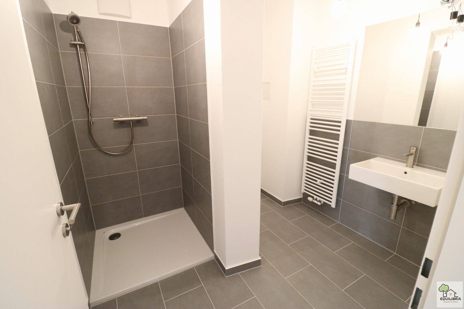 Modernes Badezimmer mit grauen Fliesen, ebenerdiger Dusche und Handtuchheizkörper.