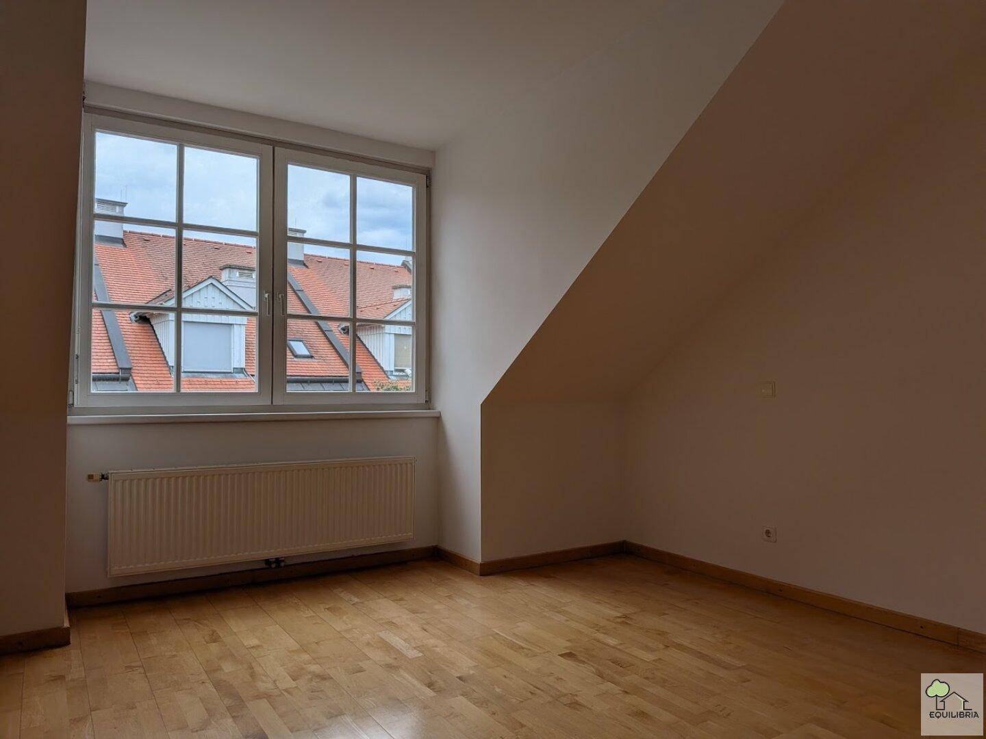 Helles Zimmer im Dachgeschoss mit schrägen Wänden, Holzparkett und großem Fenster.