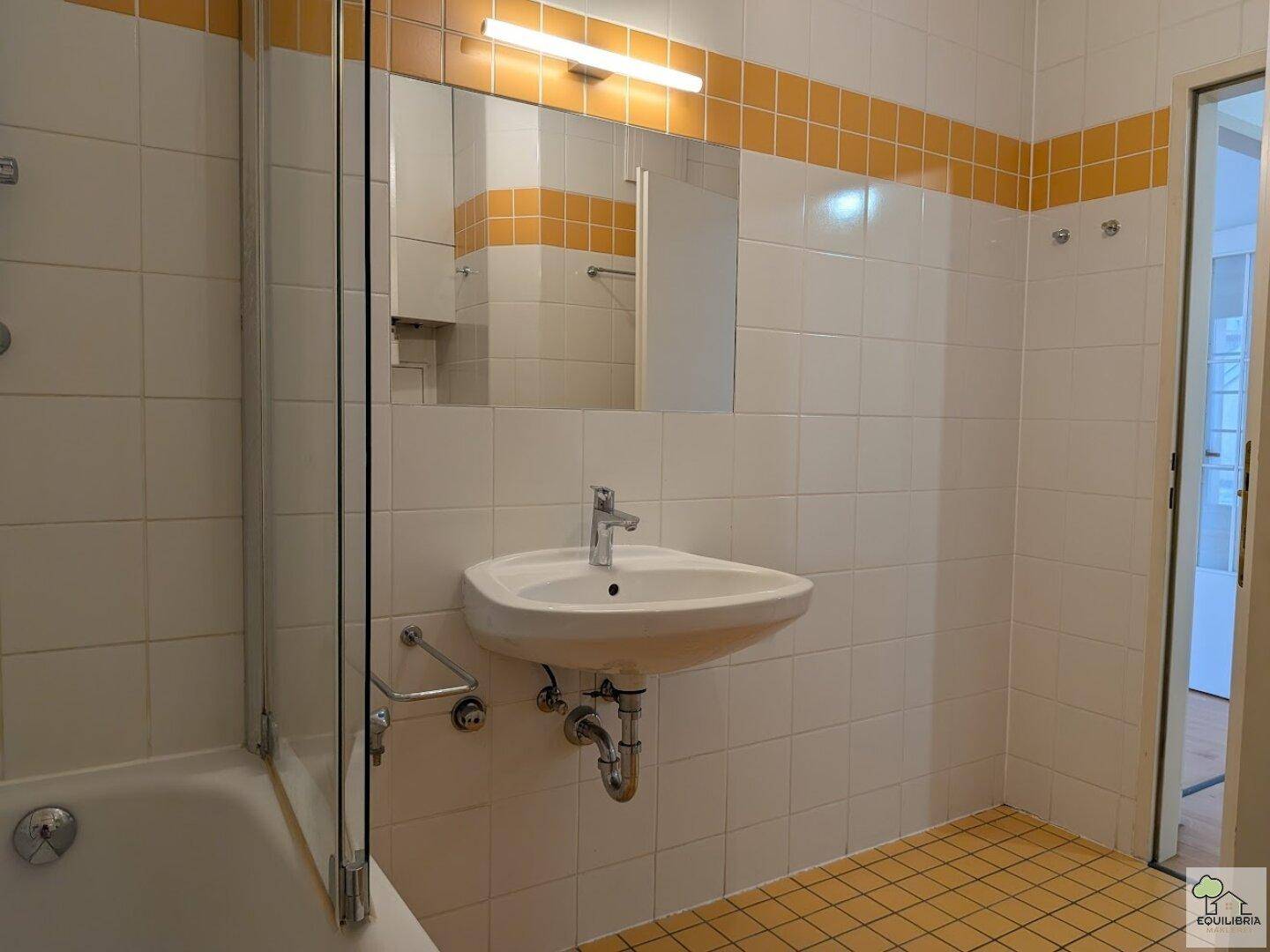 Badezimmer mit Badewanne, Waschbecken und Spiegel, gefliest in Weiß und Gelb.