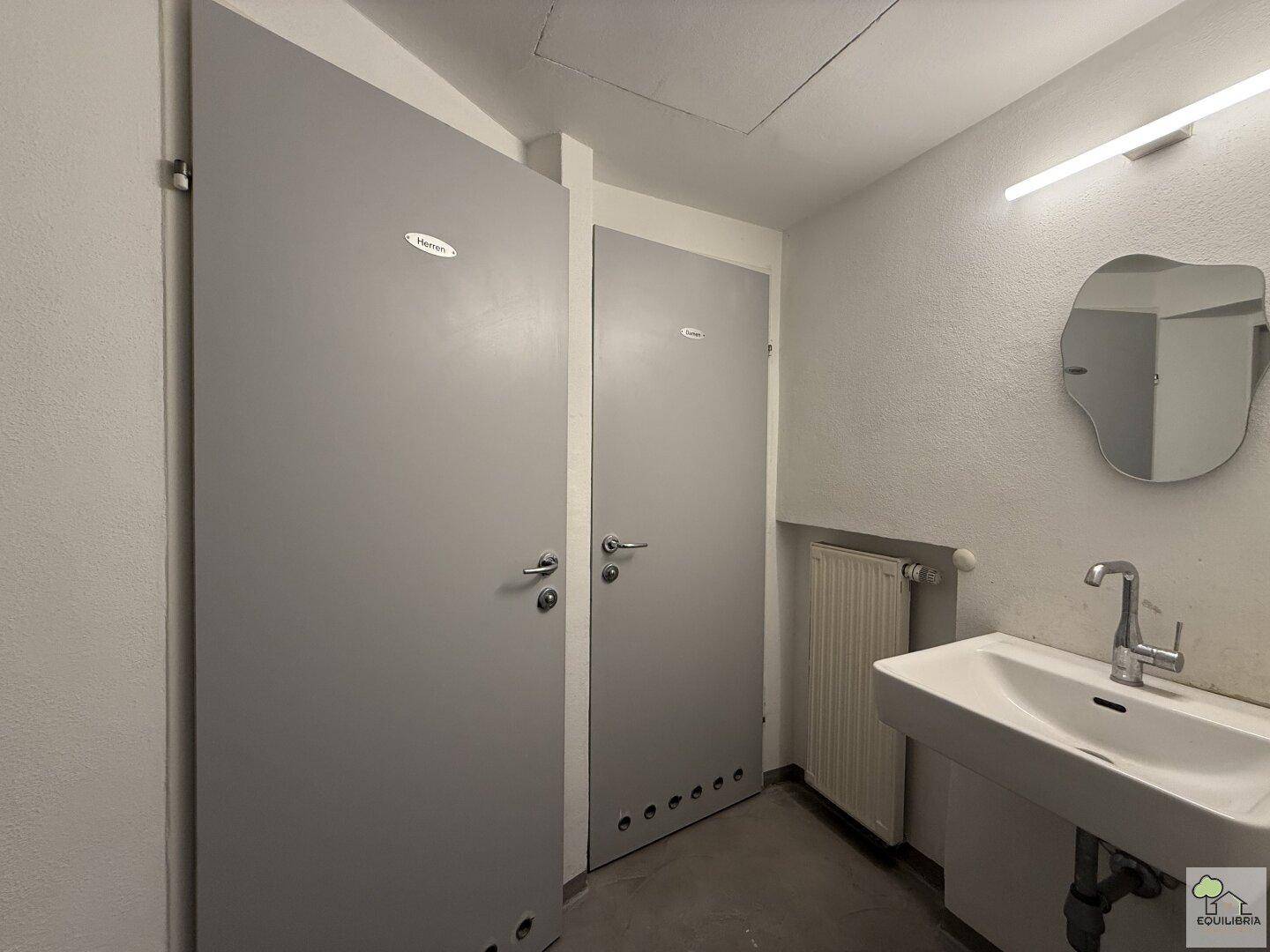 Sanitärbereich mit mehreren Toilettenkabinen, Waschbecken und Spiegel, funktional eingerichtet.