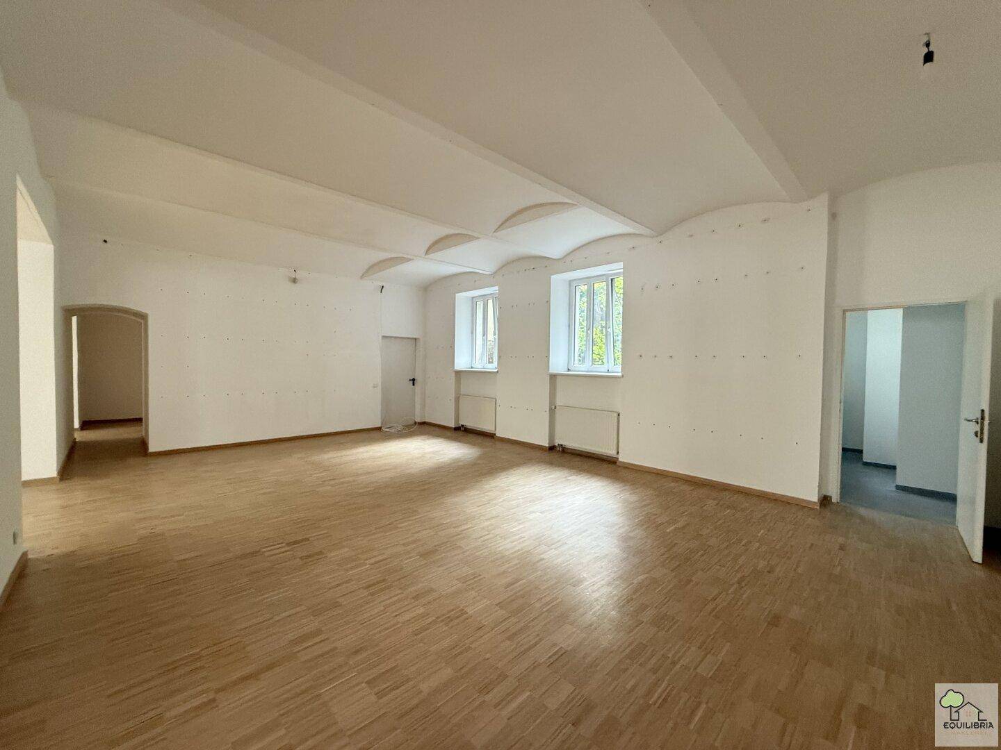 Geräumiger, leerer Wohnbereich mit Holzboden und hohen Decken, durchflutet von natürlichem Licht.