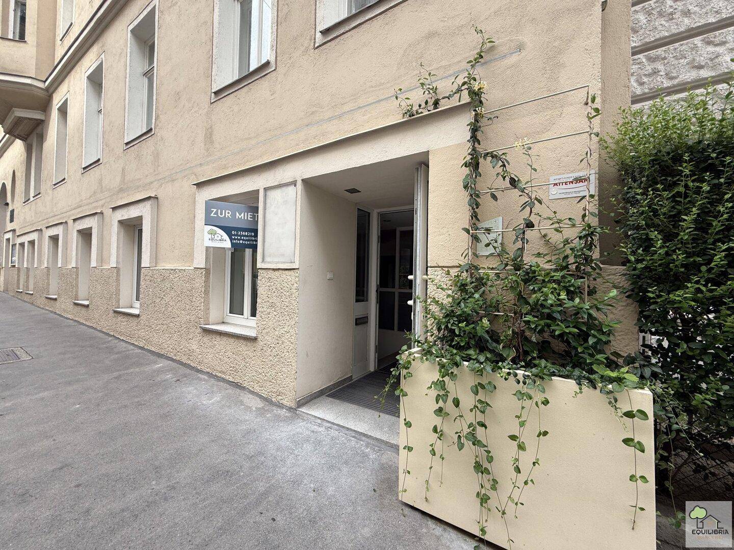 Eingangsbereich eines Mehrfamilienhauses mit "Zu vermieten"-Schild und Pflanzenkübel.