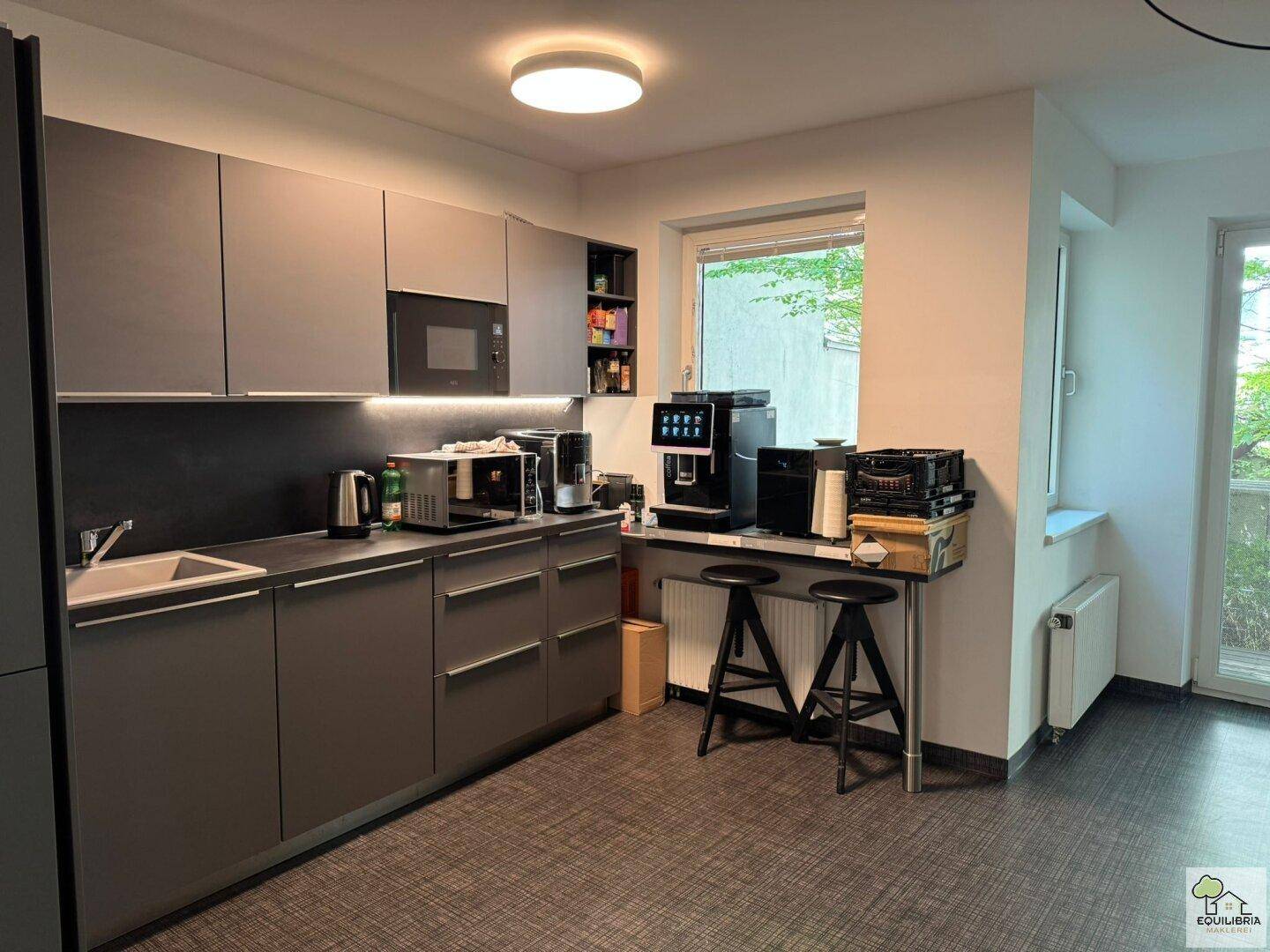 Moderne Büroküche mit grauen Schränken, integrierten Geräten und einer kleinen Bar mit Hockern.