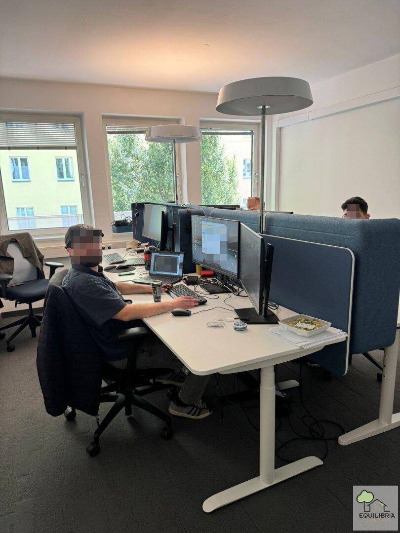 Heller Großraumbüro mit mehreren Arbeitsplätzen, modernen Schreibtischen und Computermonitoren.