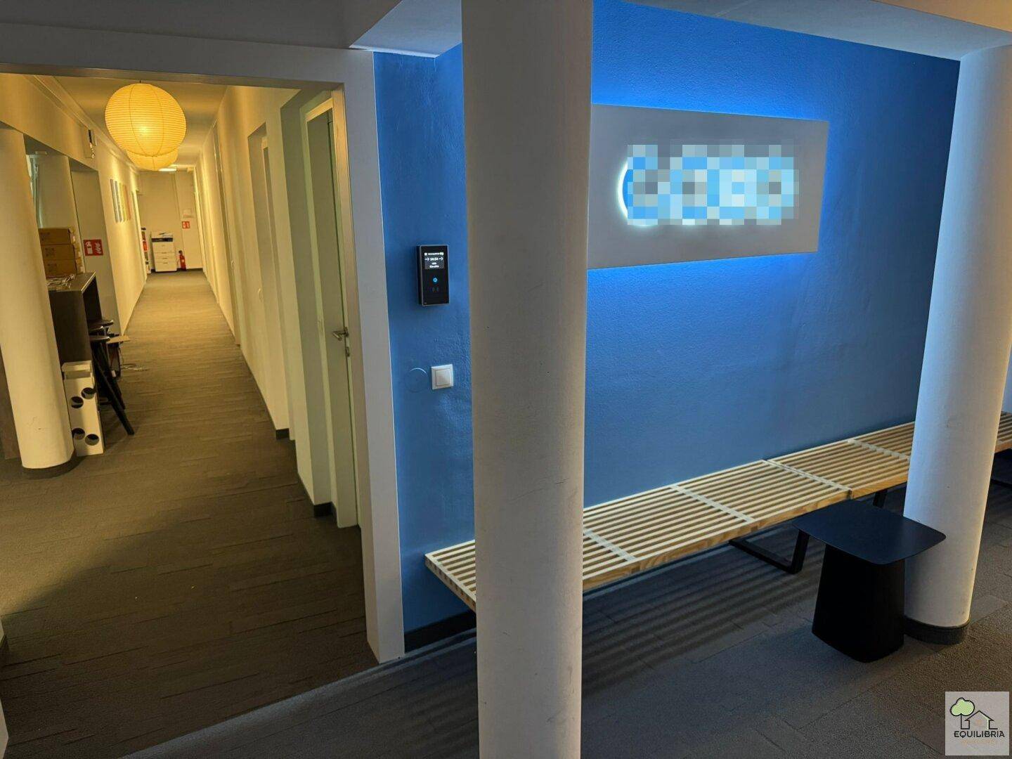 Langer Flur mit blauer Akzentwand, Sitzbank und moderner Beleuchtung in einem Bürogebäude.