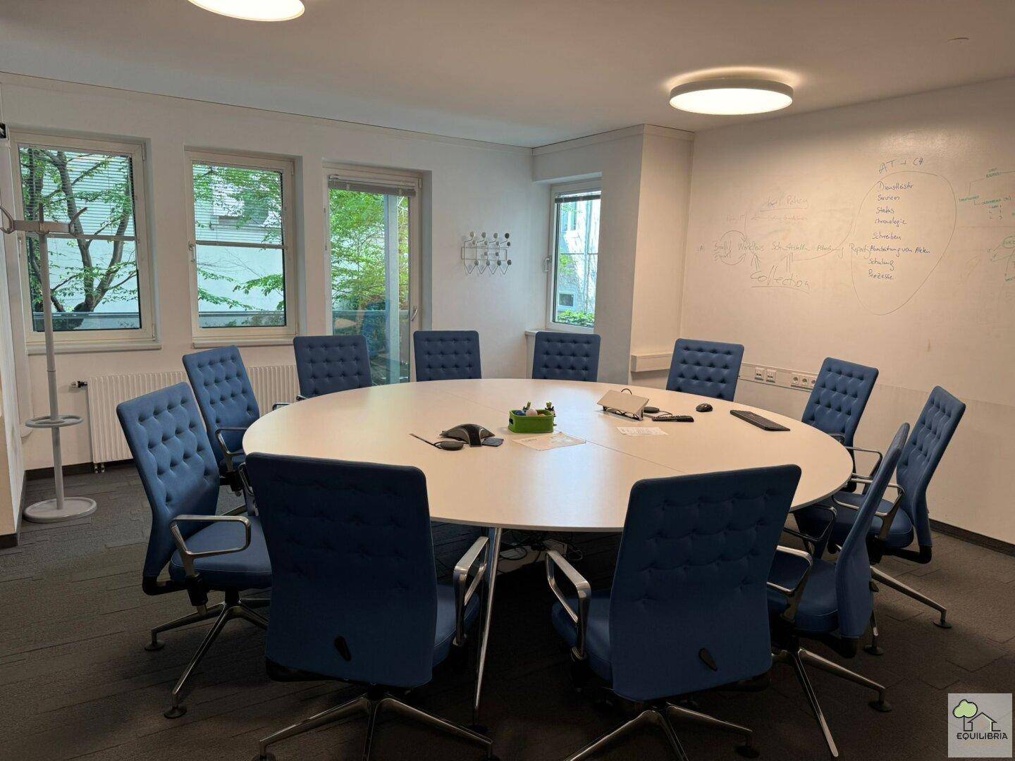 Geräumiger Konferenzraum mit großem ovalen Tisch, blauen Stühlen und Fenstern für Tageslicht.