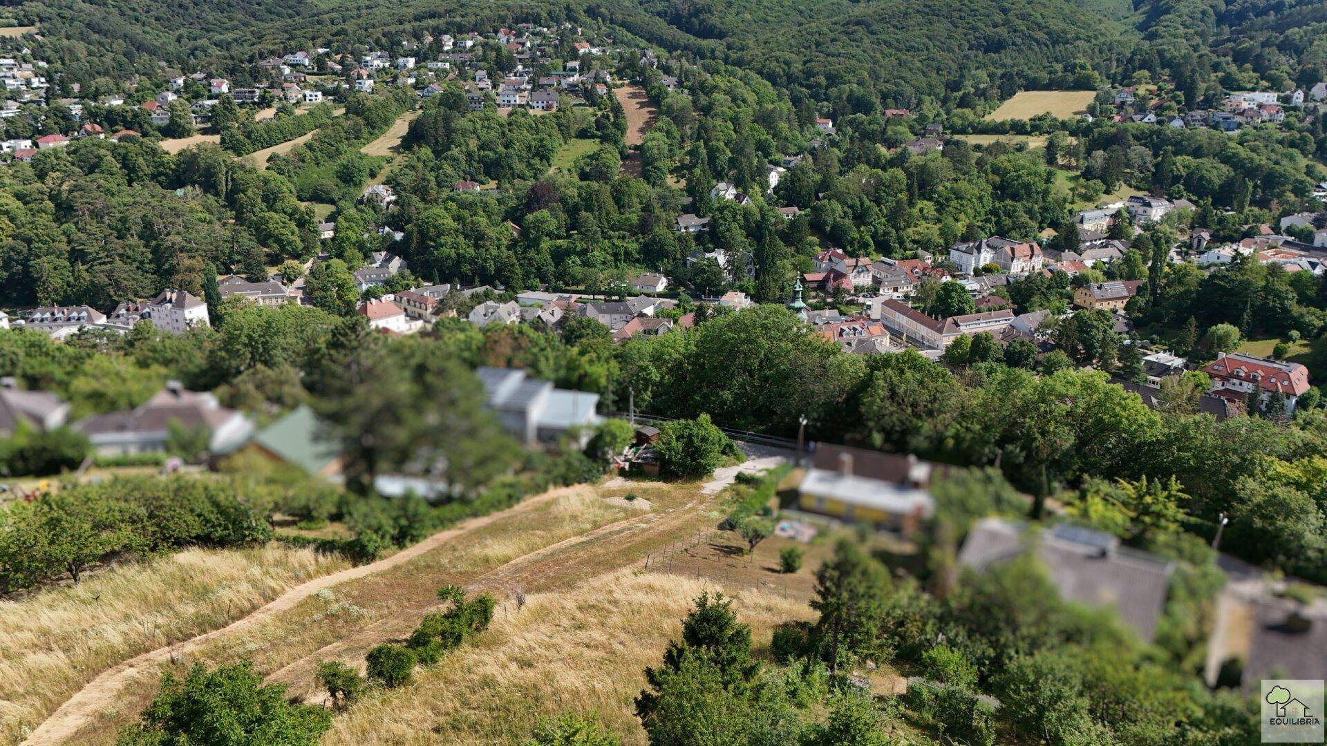 Panoramablick aus der Vogelperspektive auf eine malerische Ortschaft, eingebettet in eine grüne, bewaldete Hügellandschaft.