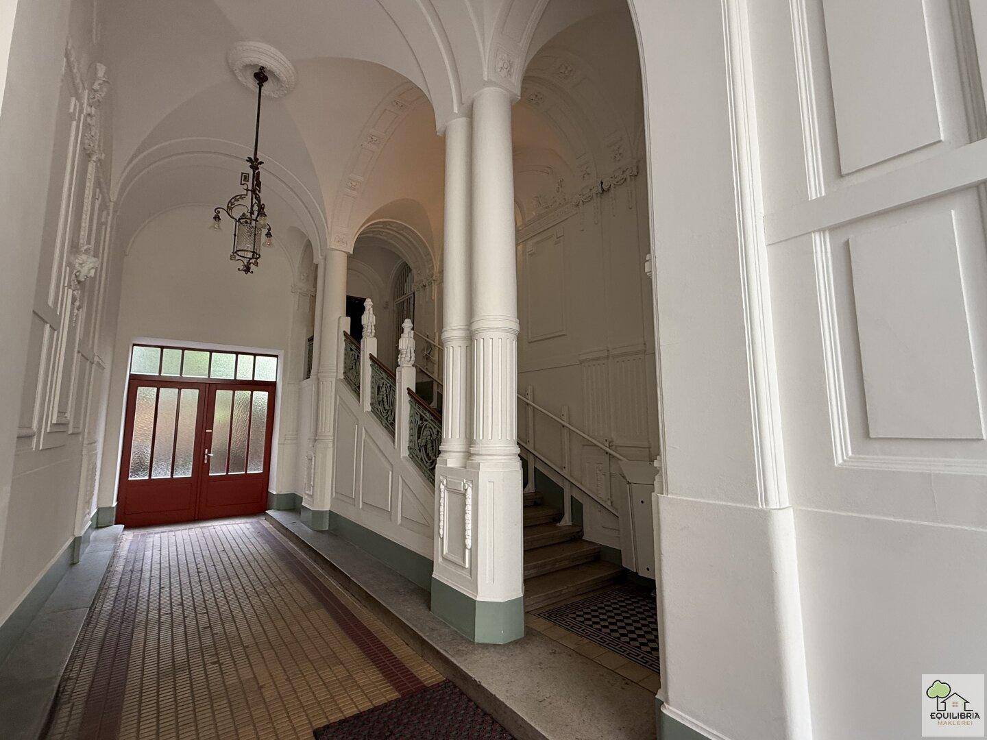Großzügiger Eingangsbereich eines Altbaus mit hohen Gewölbedecken, Säulen und einer eleganten Treppe.