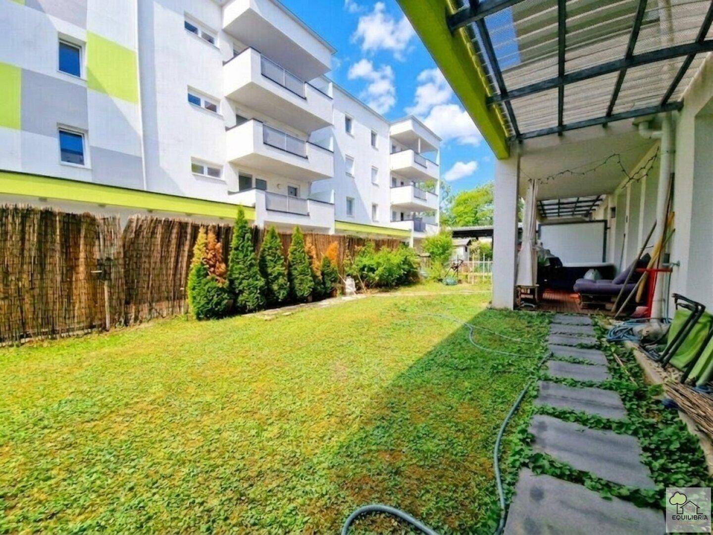 Weitläufiger Garten mit Rasenfläche, Steinpfad und überdachtem Terrassenbereich am modernen Gebäude.