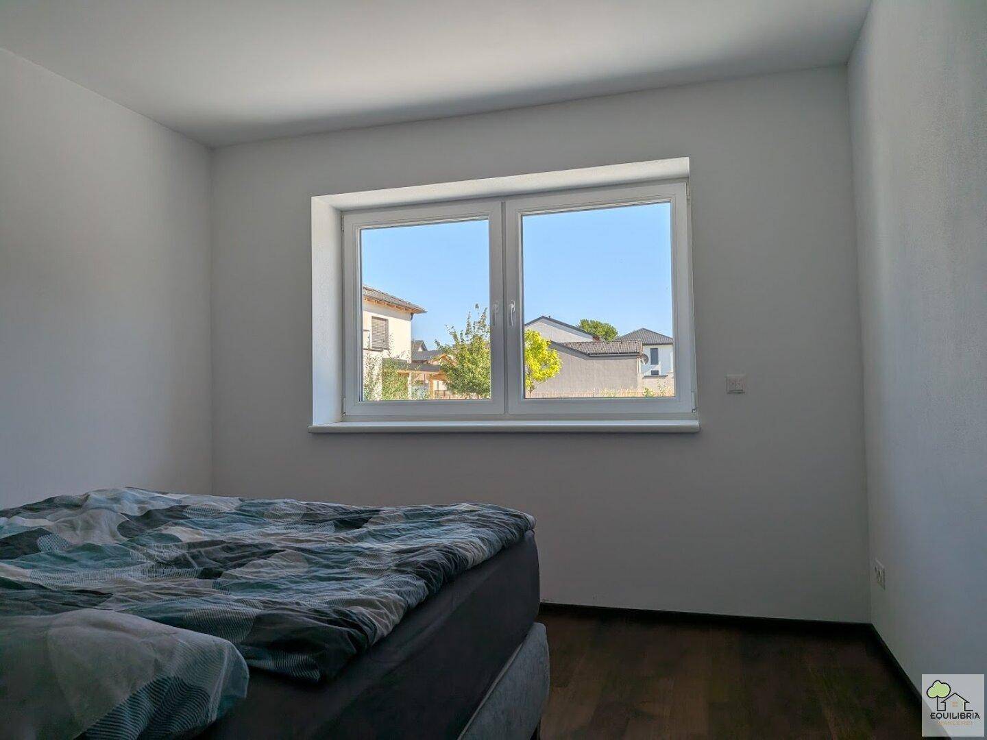 Schlafzimmer mit Holzboden und großem Fenster, das viel Tageslicht hereinlässt.